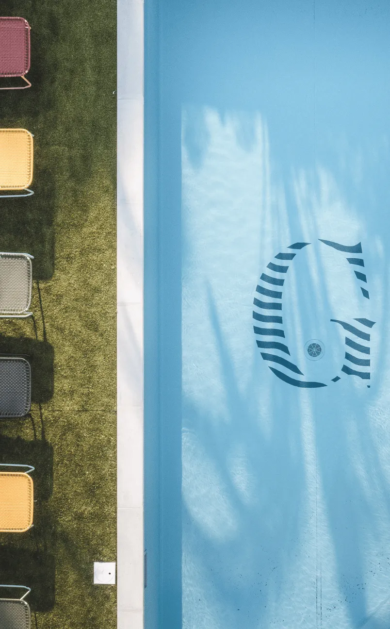 Aerial view of a clean blue swimming pool with a stylized 'G' logo at the bottom and colorful chairs lined up on green grass beside it.