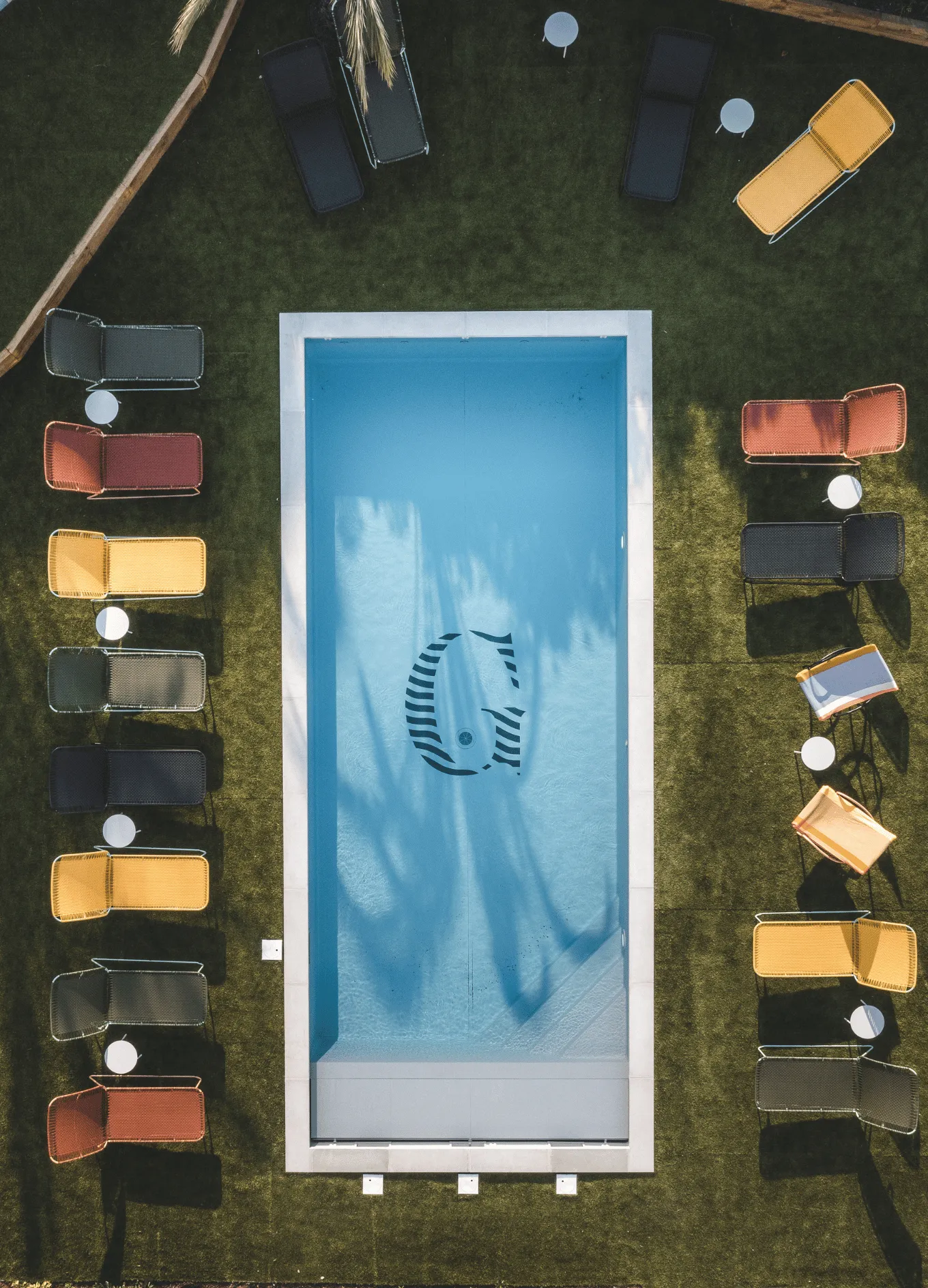 Overhead view of a rectangular blue pool with a large G logo at the bottom, surrounded by green grass and colorful lounge chairs with small round tables.