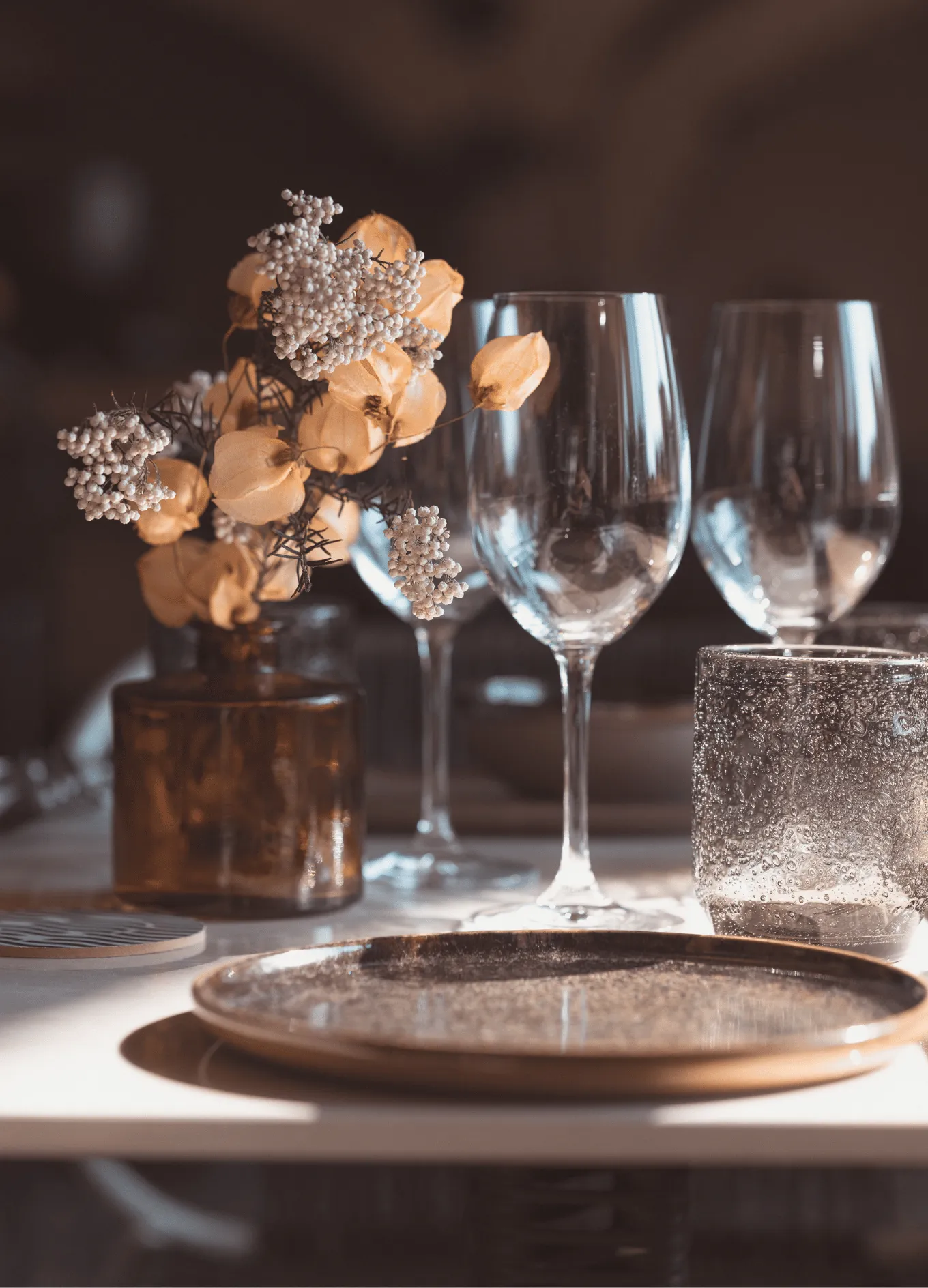 Close-up of a table setting with empty wine glasses, a textured drinking glass, a brown vase with dried flowers, and a ceramic plate.