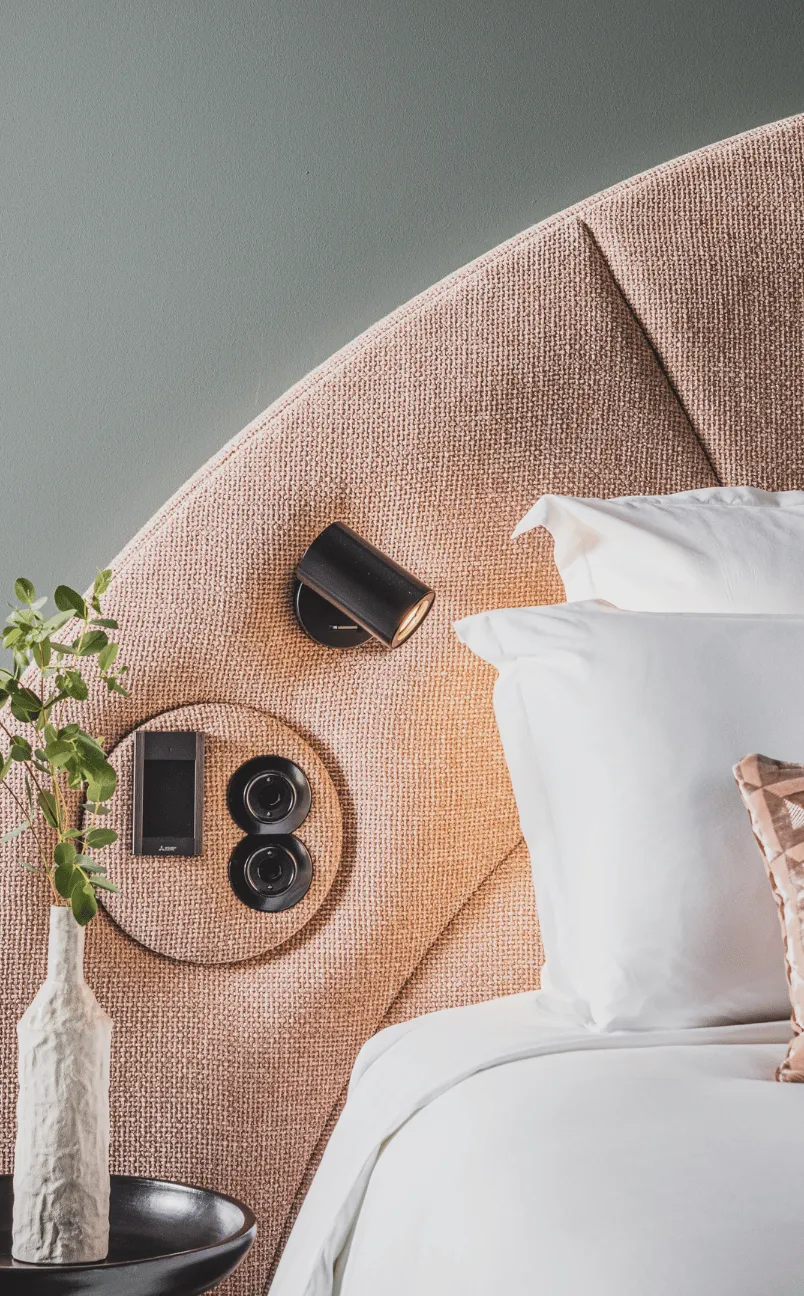Close-up of a pink textured hotel bed headboard with a small black wall light, control panel, and a vase with green leaves on a side table.