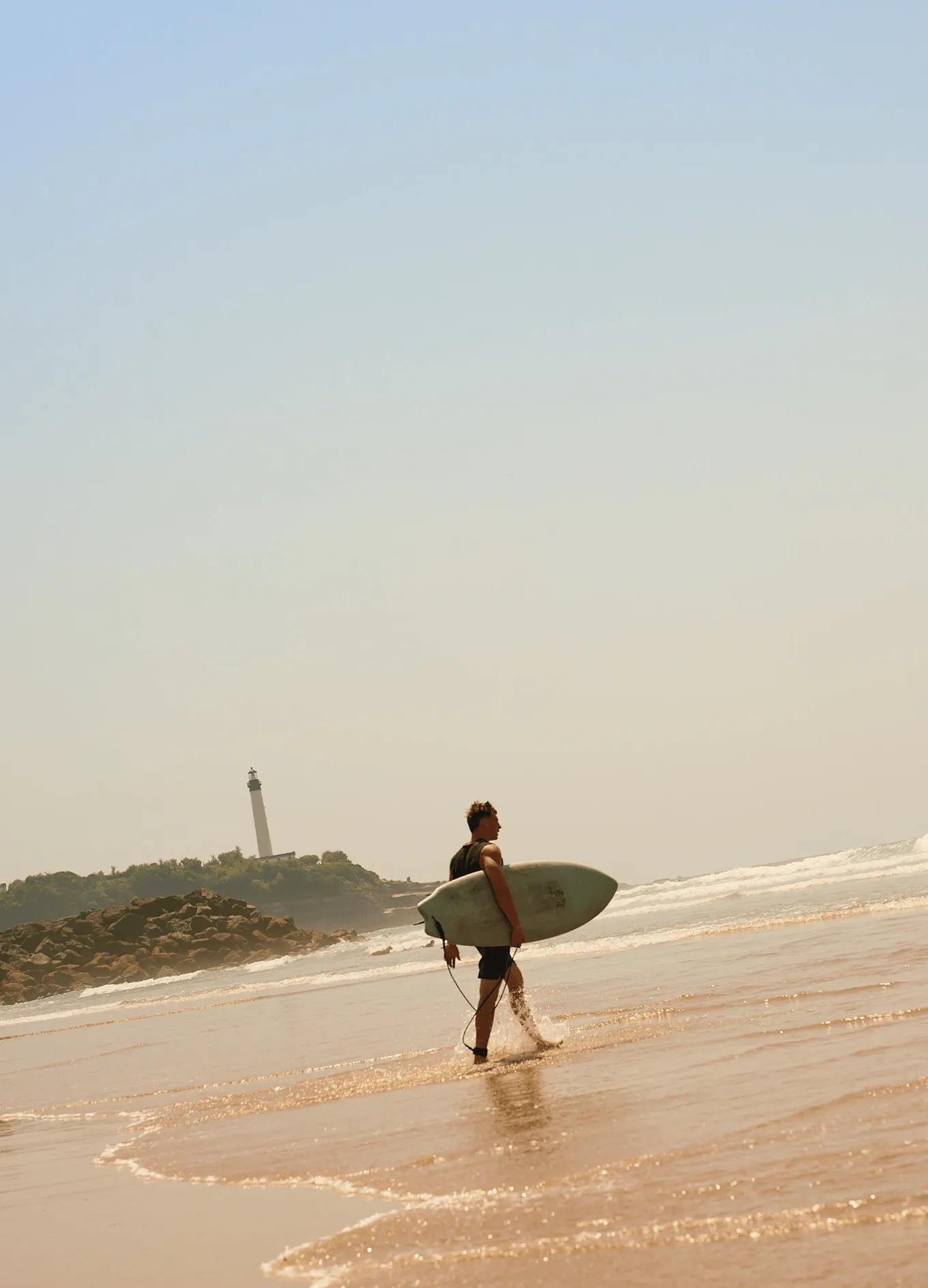Surfer walking through shallow ocean water carrying a surfboard with a lighthouse and rocky coastline in the background.