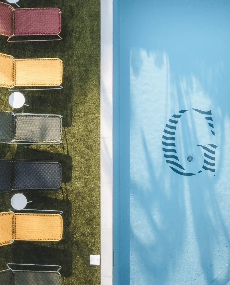 Top-down view of colorful lounge chairs with small round tables on a grass lawn next to a blue pool with a large letter G at the bottom.