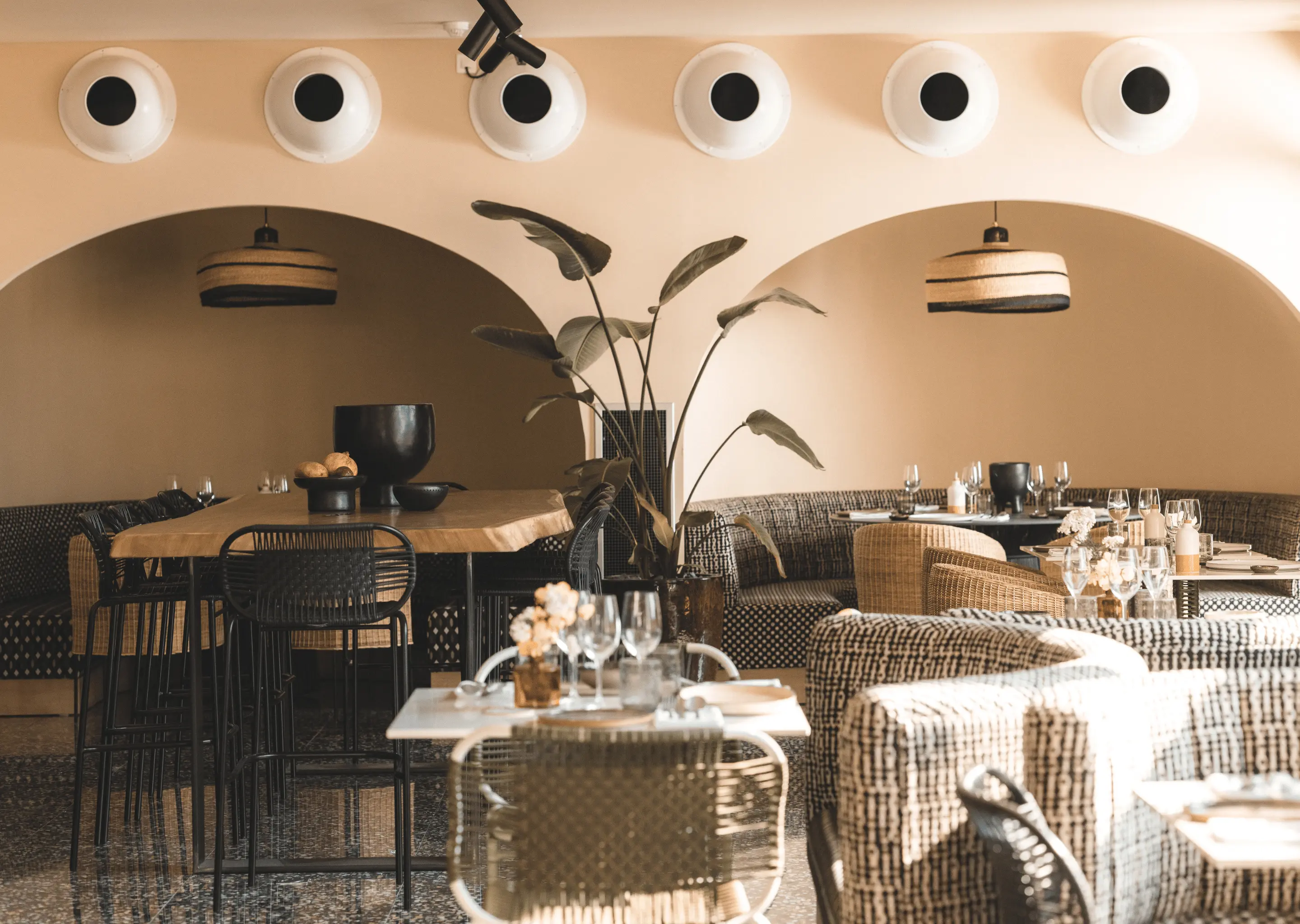 Cozy restaurant interior with curved banquette seating, wooden tables, woven chairs, and potted tropical plant under beige arches.