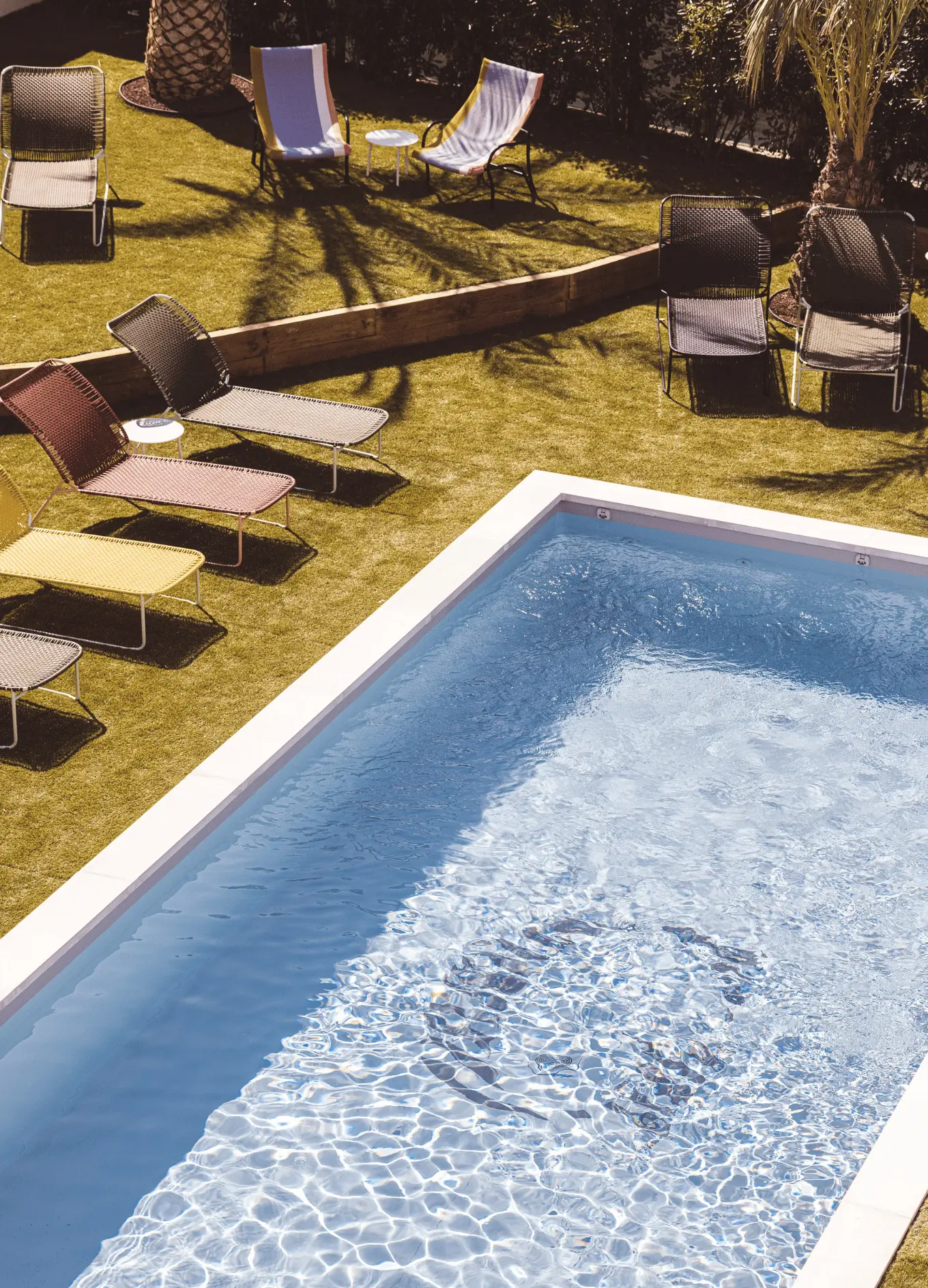 Small outdoor pool with clear water next to green lawn with various colored lounge chairs and palm trees casting shadows.
