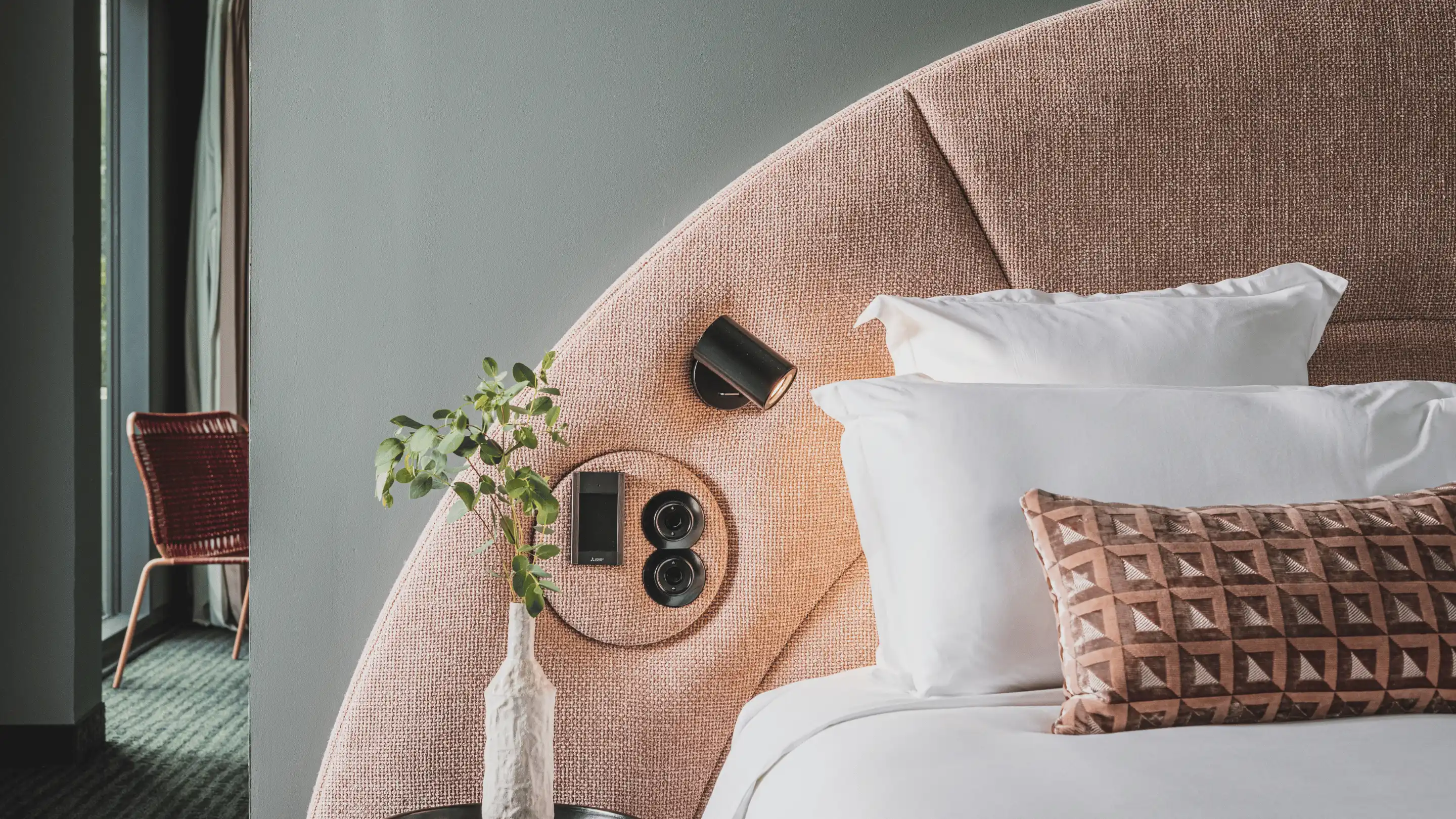 Close-up of a bed with white pillows, a patterned brown cushion, beige textured headboard with built-in black reading light and controls, and a vase with green leaves on a side table.