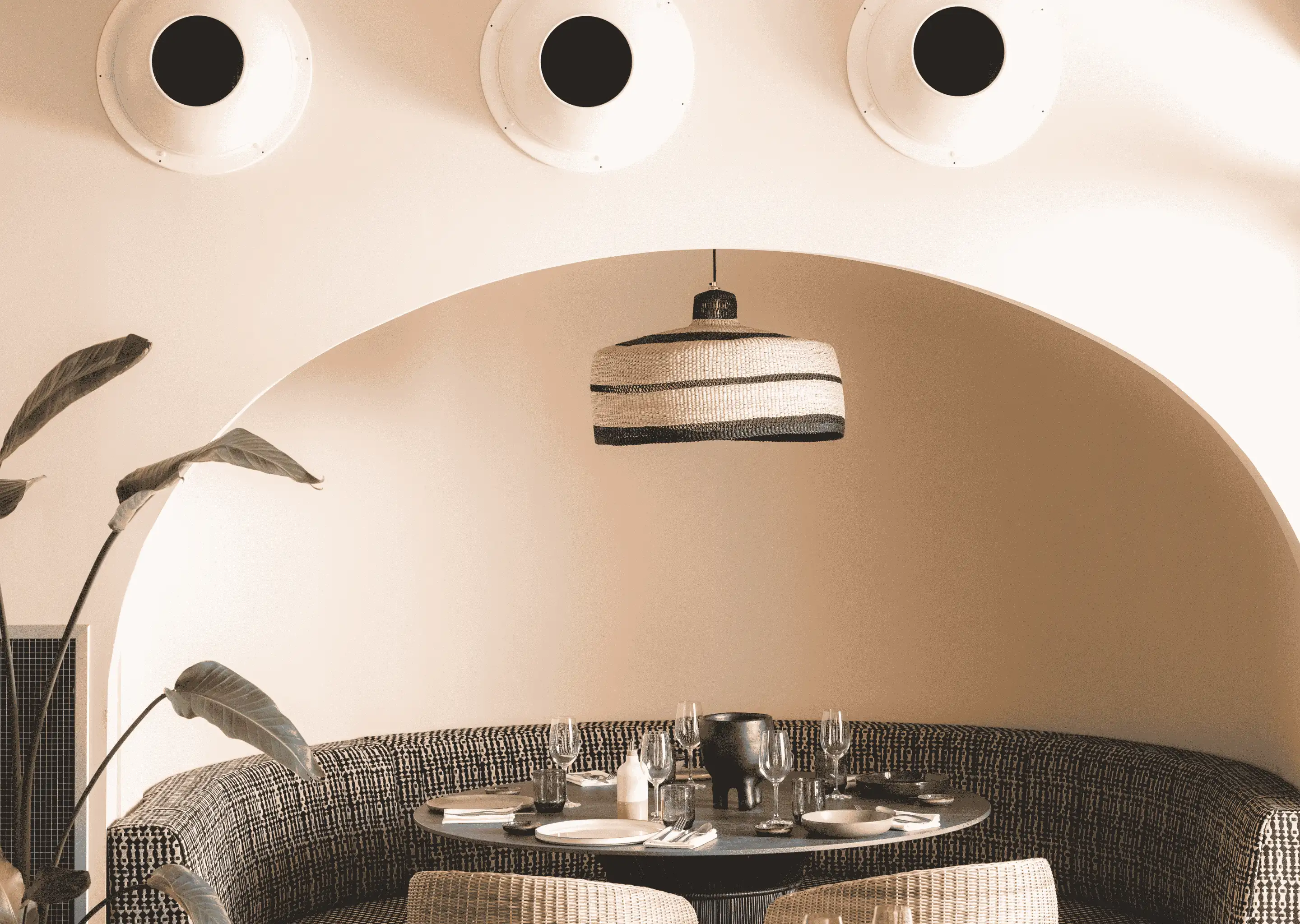 Curved dining booth with patterned upholstery around a round table set with plates and glasses under a woven pendant light in a minimalist dining area.