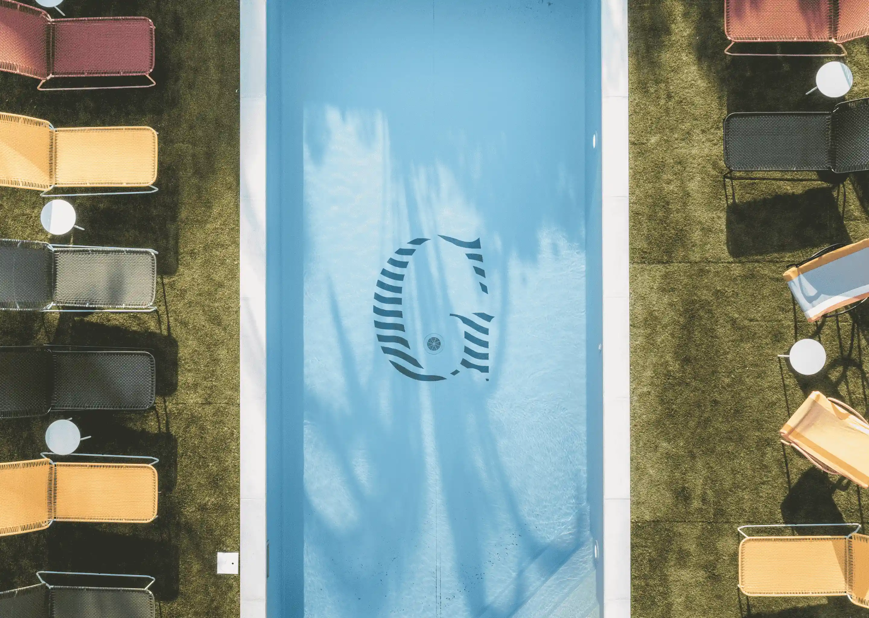 Aerial view of an empty rectangular swimming pool with a stylized letter G at the bottom, surrounded by lounge chairs and small round tables on grass.