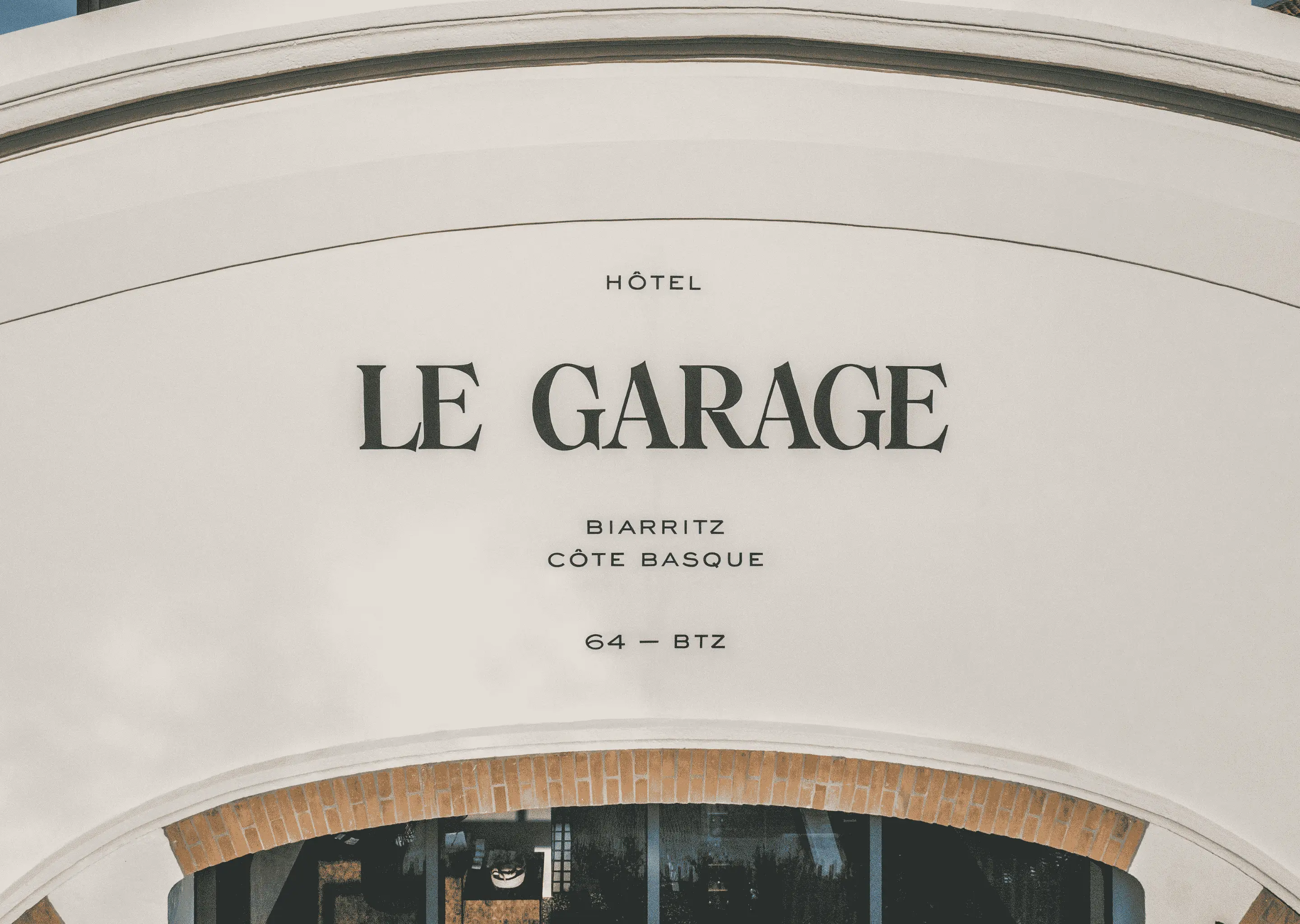 White arched wall with black text reading Hôtel Le Garage Biarritz Côte Basque 64 - BTZ above a brick-lined window.