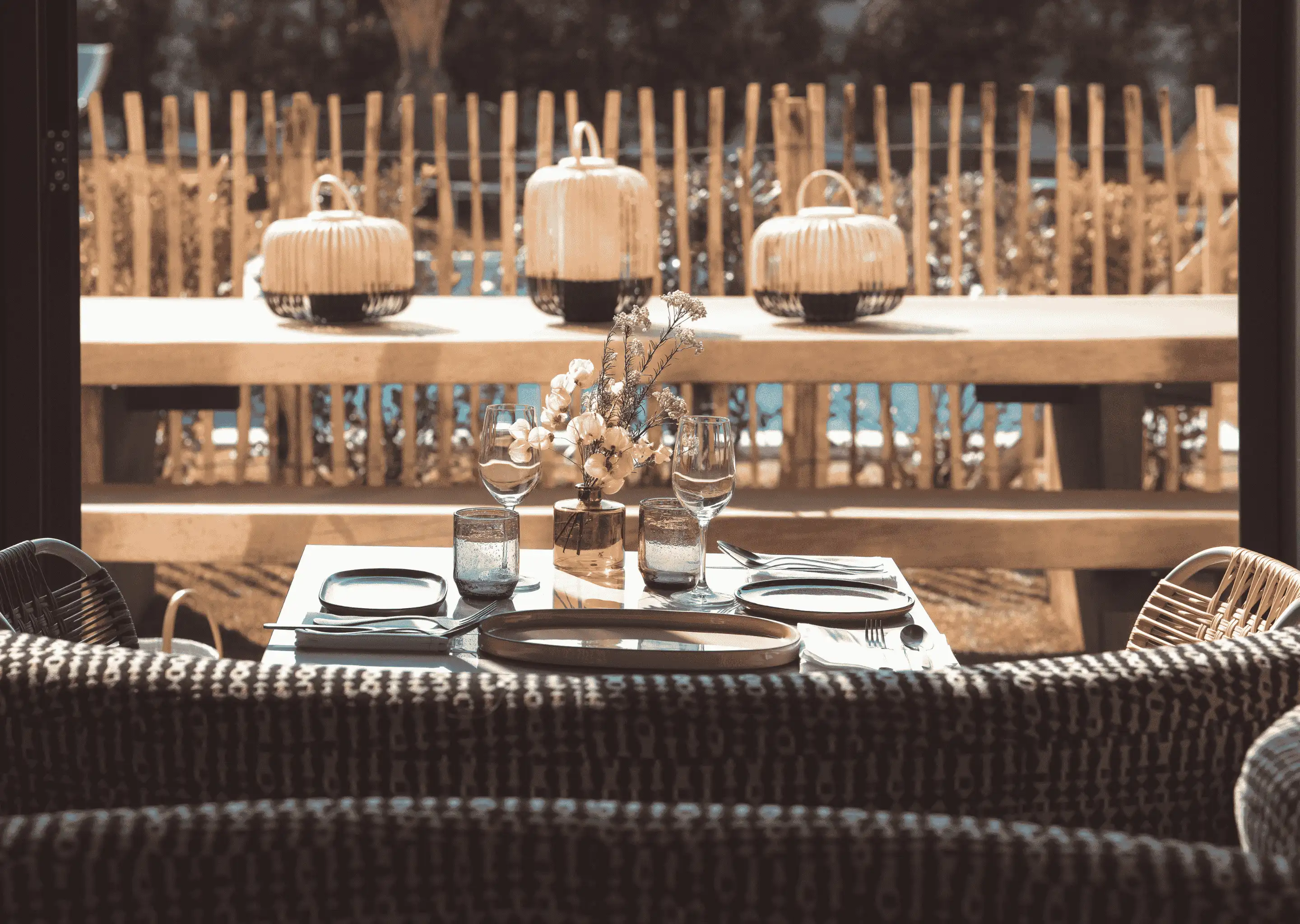 Dining table set with glassware, plates, cutlery, and a small floral centerpiece, with wicker chairs and lanterns on a bench behind it.
