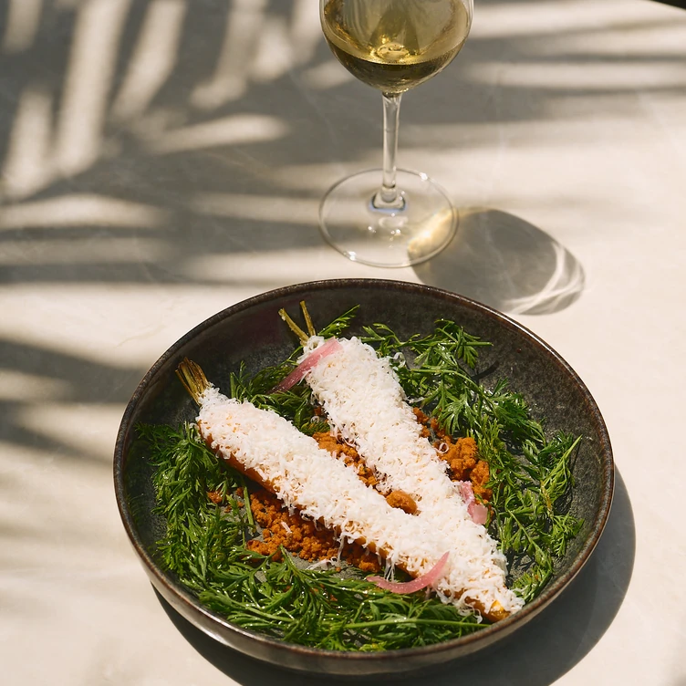 Two whole carrots covered with grated cheese served on a bed of green carrot tops and crumbs in a dark bowl, with a glass of white wine nearby casting a shadow.