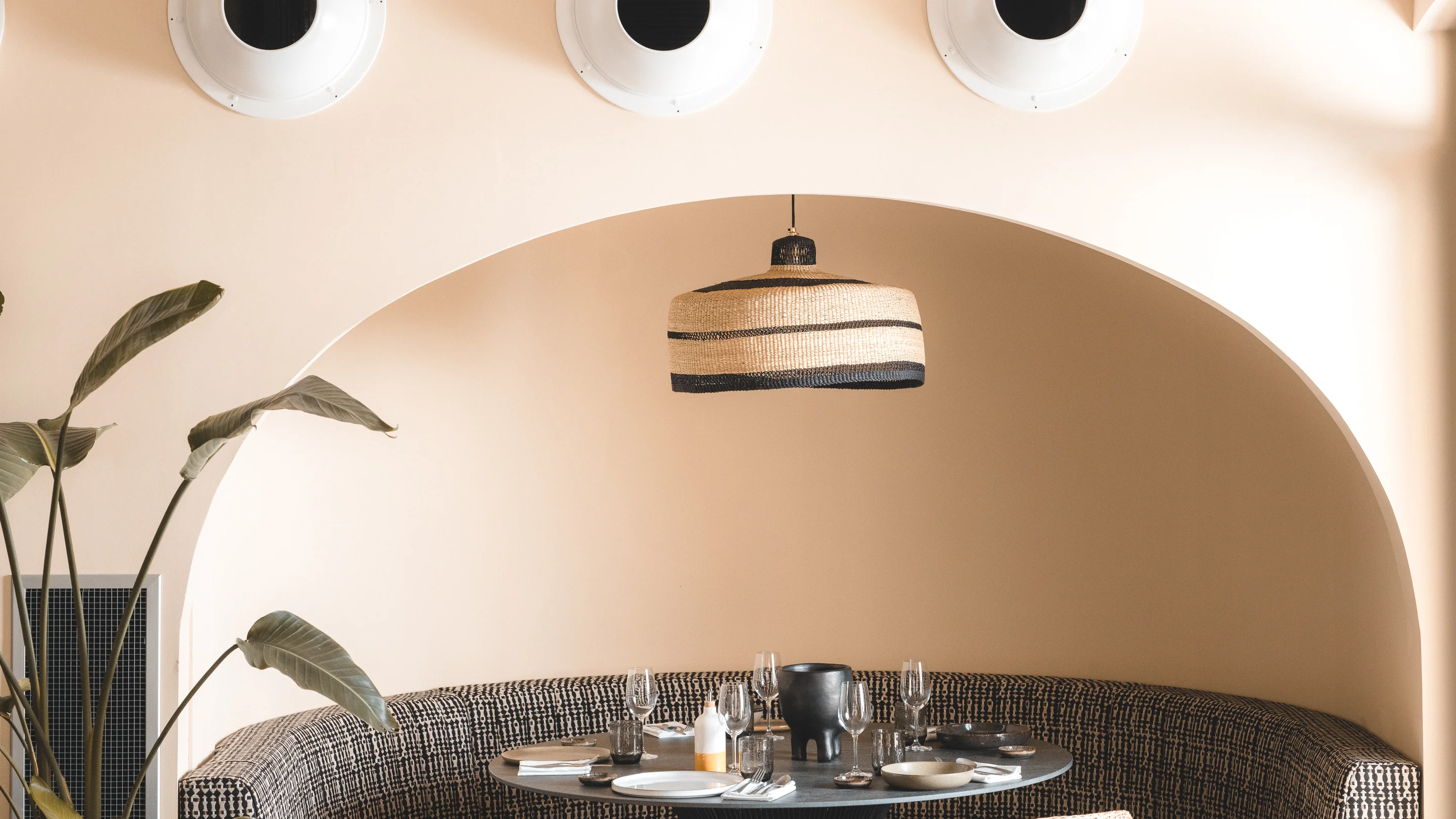 Sophisticated dining area with a curved booth, woven lighting, and a view of a lush plant at Hôtel Le Garage, Biarritz.