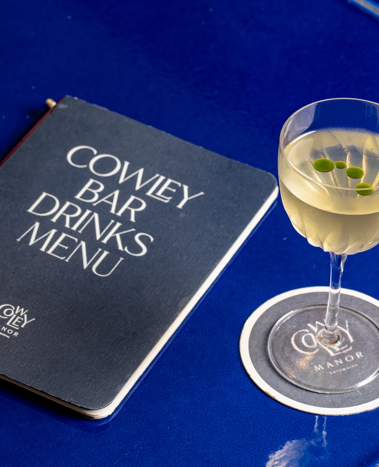 Detail of a signature cocktail and branded coaster at Cowley Manor, set on a blue bar counter.