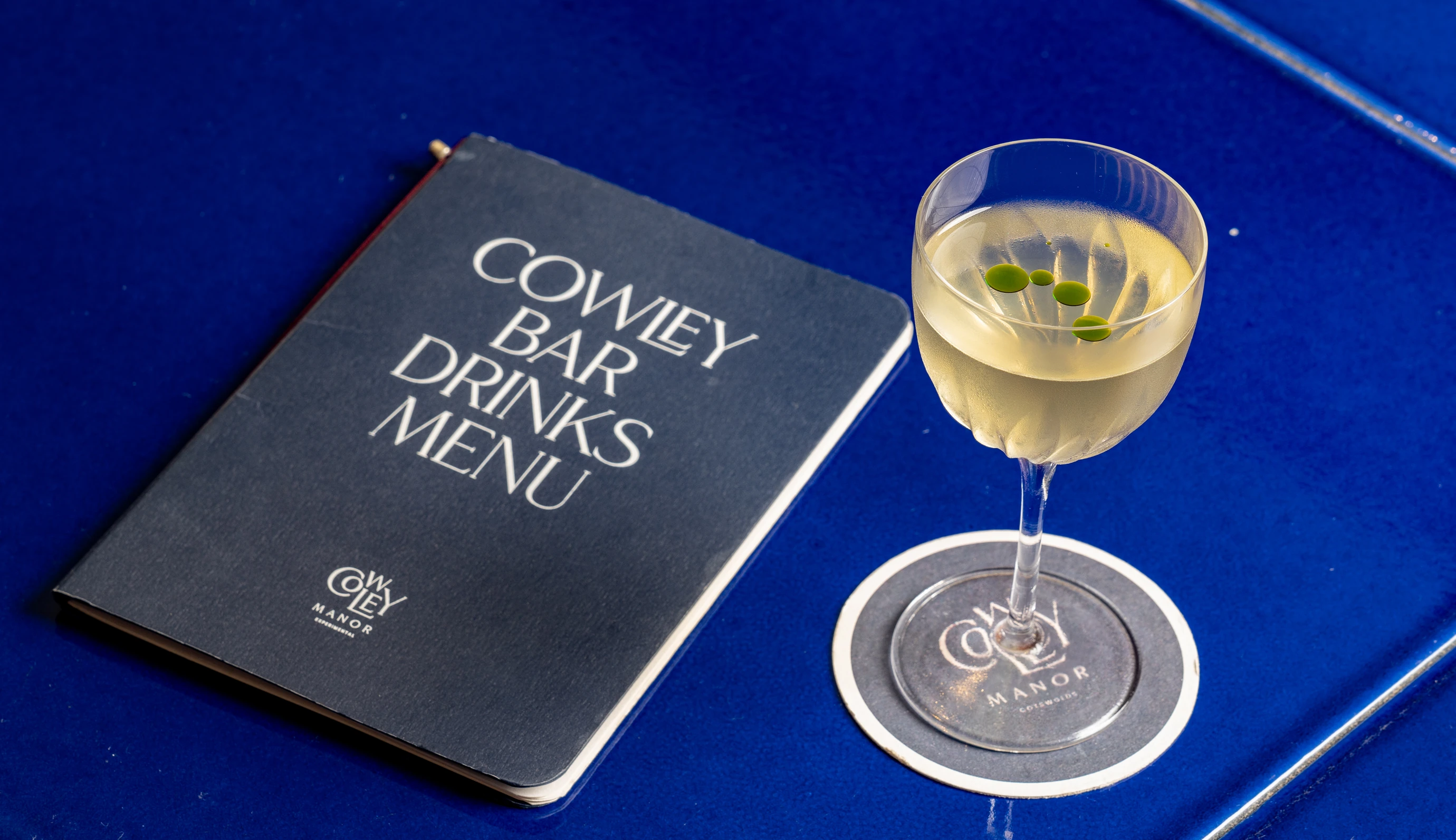 Cowley Bar drinks menu and a refined cocktail placed on a blue table at Cowley Manor, Cotswolds.