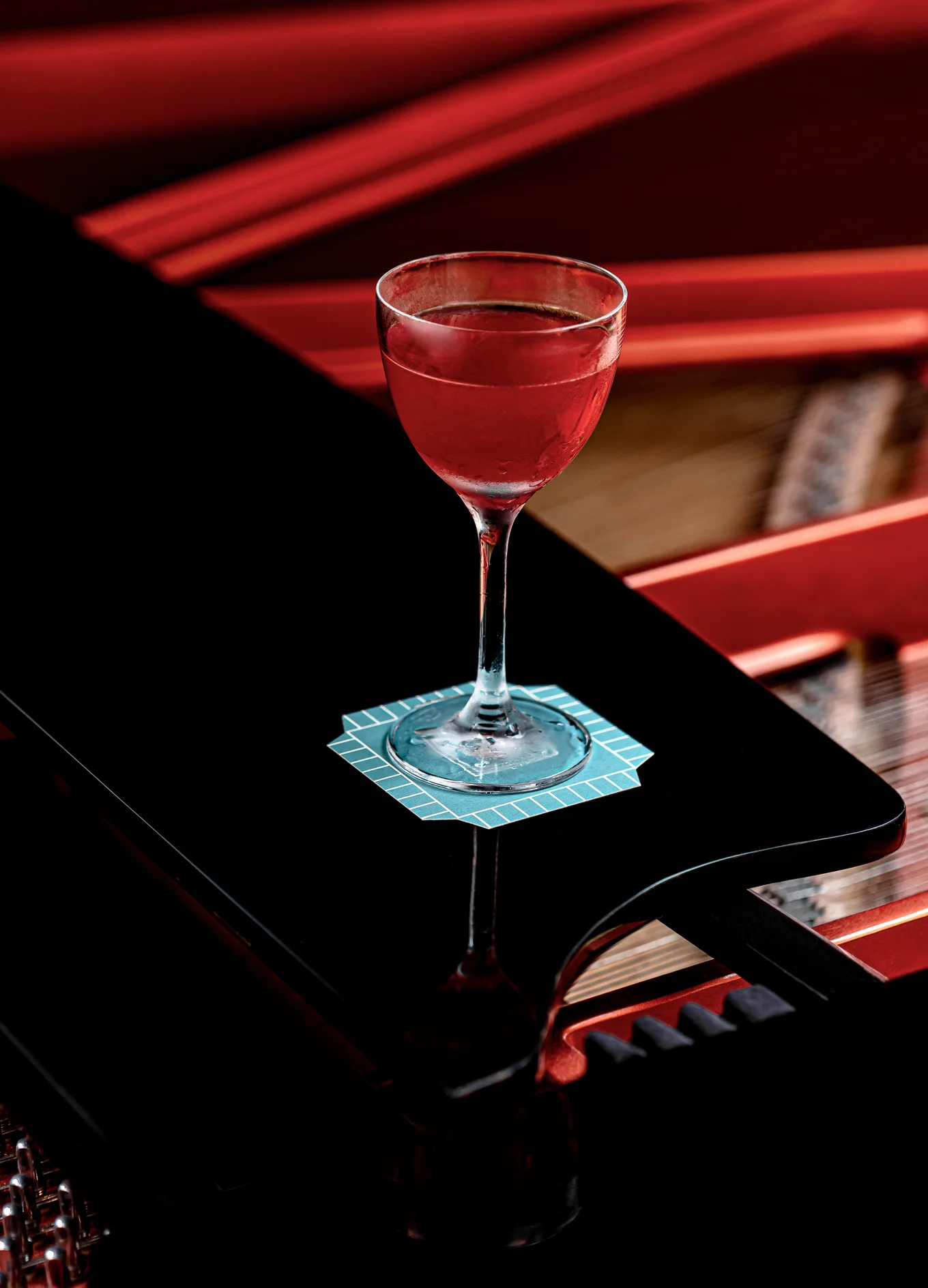 Cocktail glass with red drink on a blue coaster atop a black piano with red interior.
