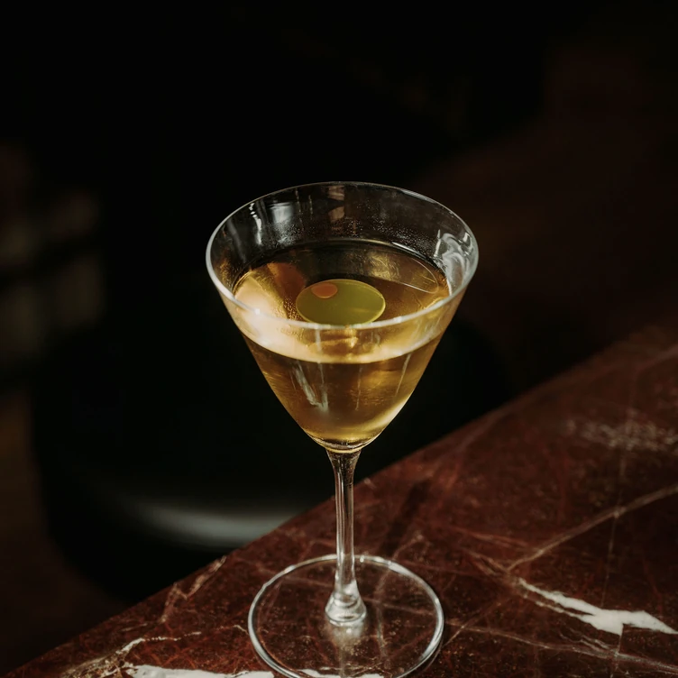 Focus on a classic Martini cocktail with an olive served on a red marble bar counter by the Experimental Group.