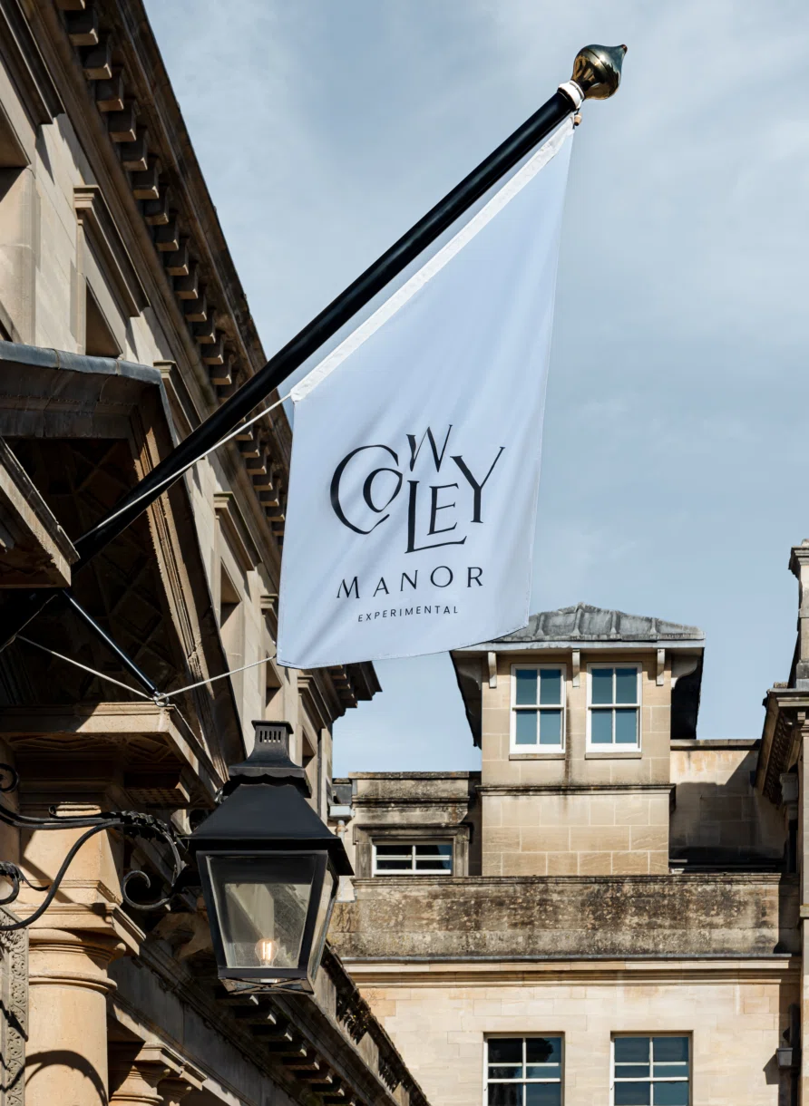 White flag with Cowley Manor Experimental logo hanging from a beige stone building next to a black lantern.