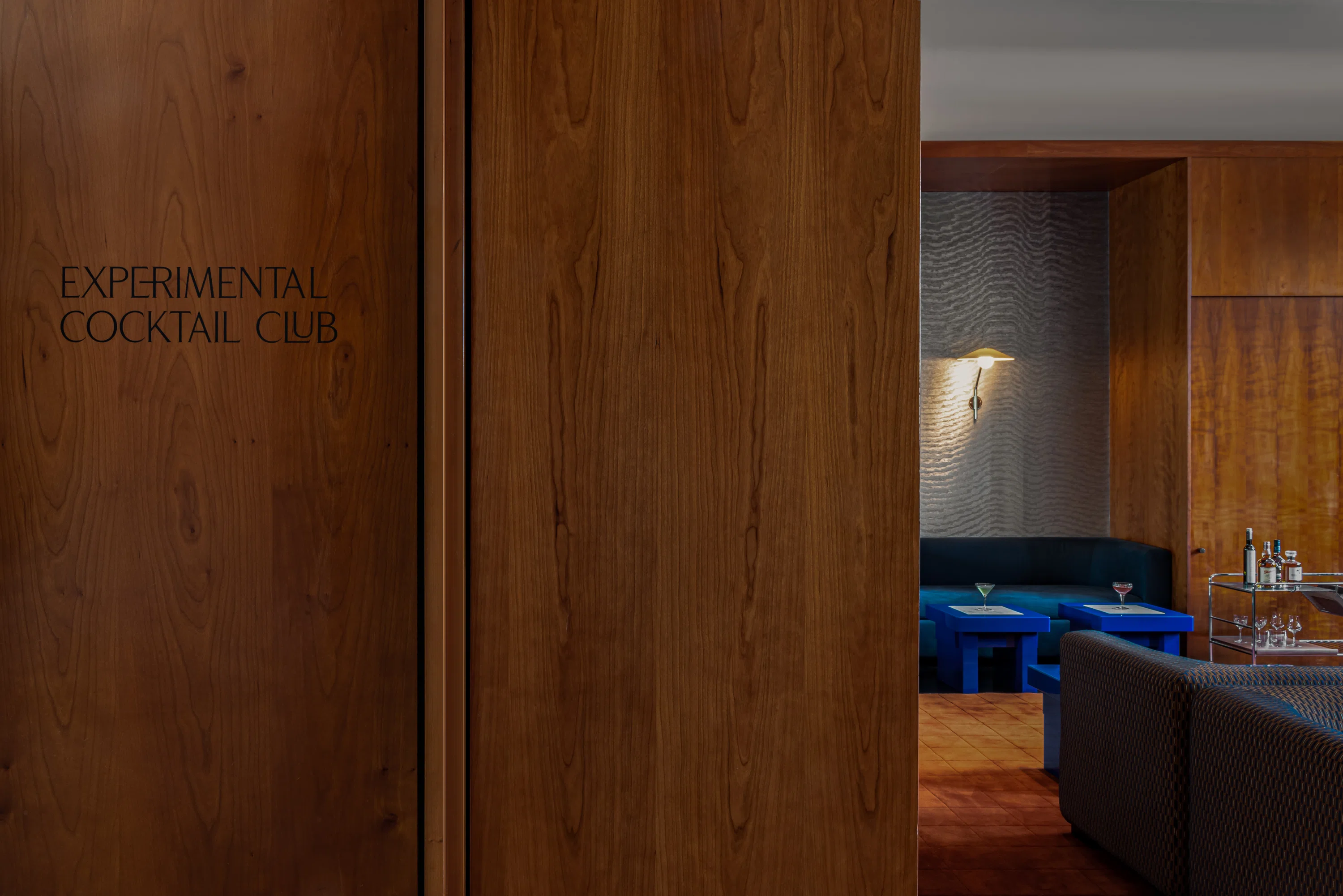 Interior of Experimental Cocktail Club with wood panels, blue tables, cocktails, and cozy seating.