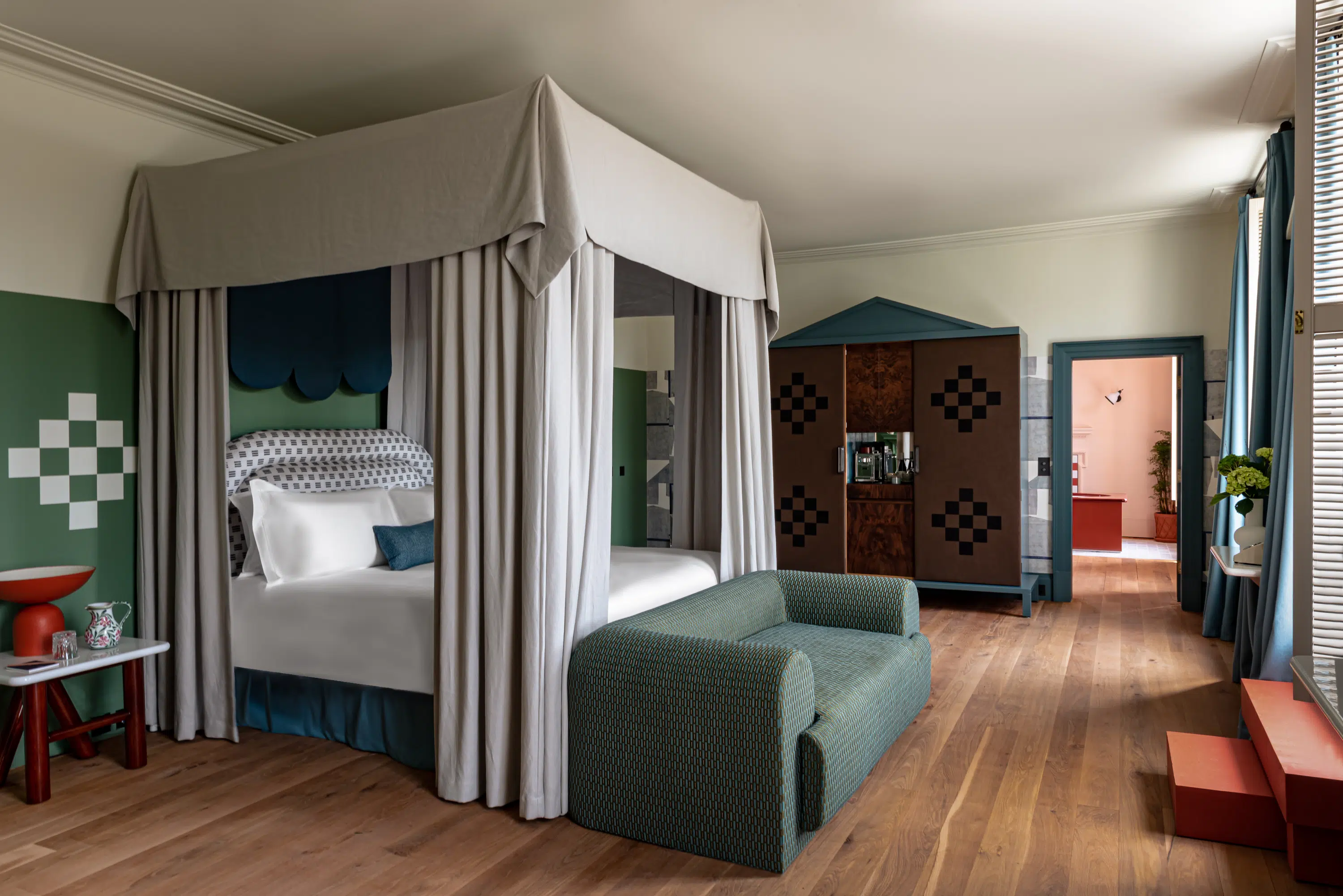 Hotel room with canopy bed, patterned green loveseat, wood floor, and blue-trimmed doorway.