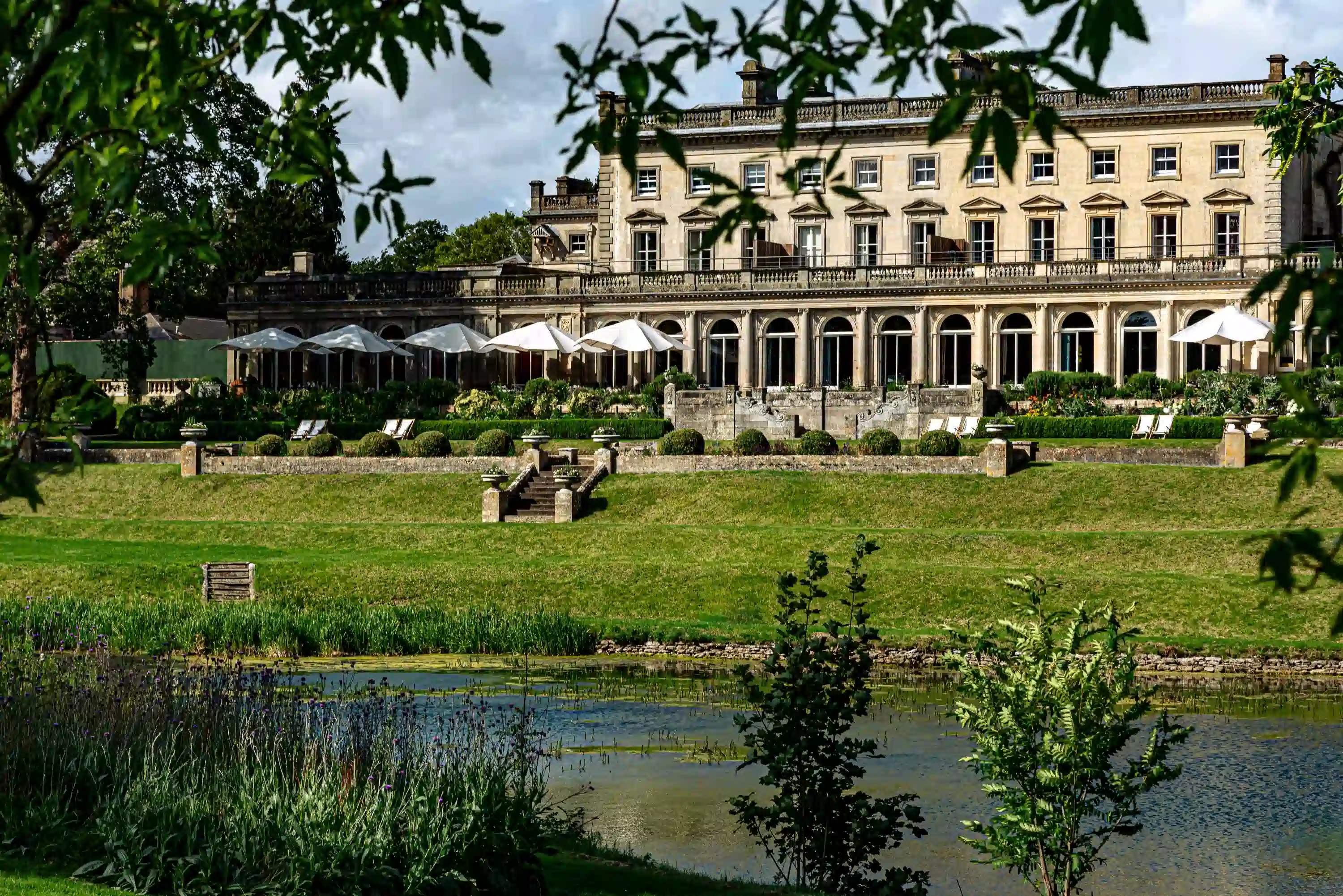 Large classic manor house with outdoor white umbrellas and a pond in front framed by leafy branches.