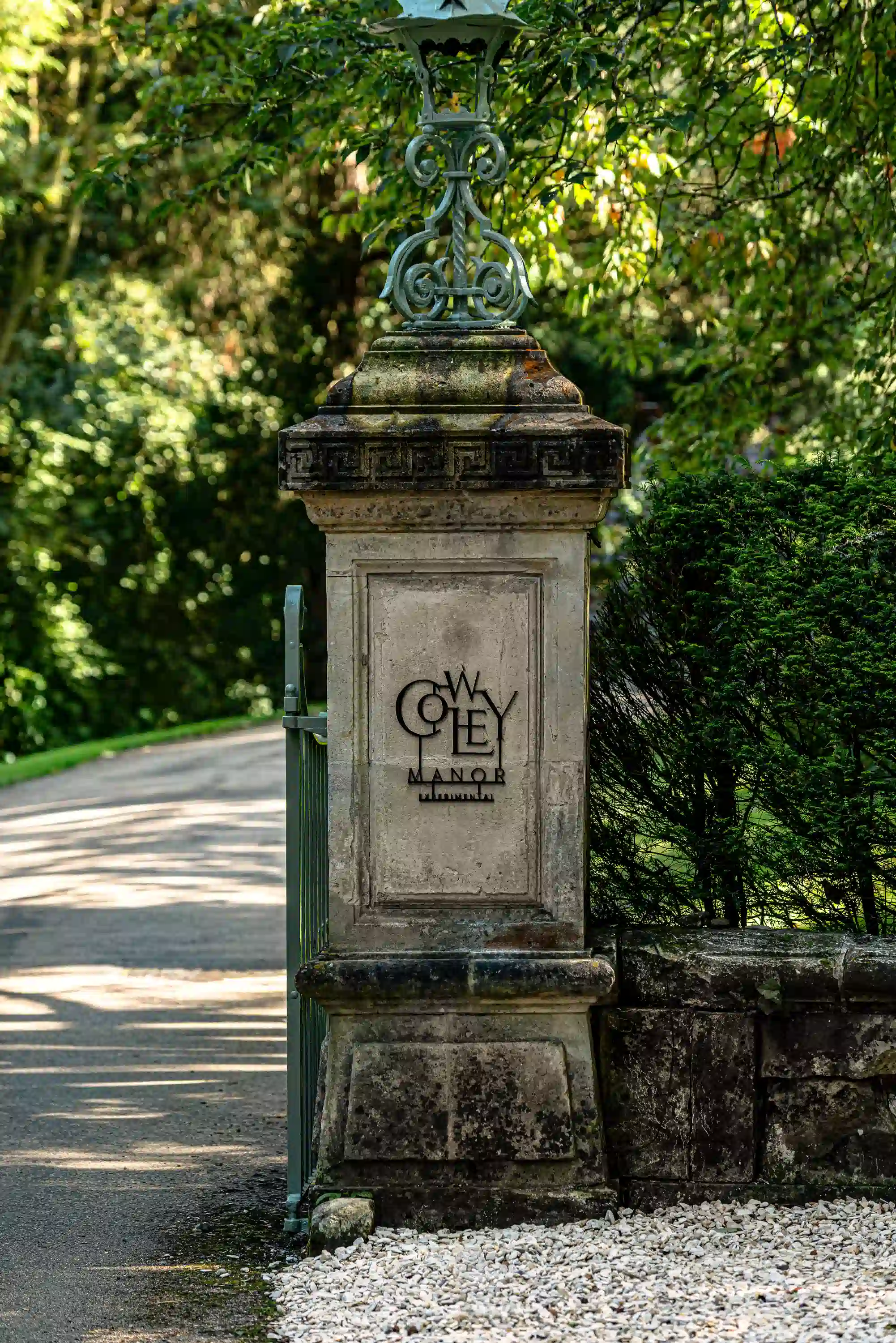 Stone pillar with Cowley Manor logo at the entrance surrounded by greenery and a driveway.