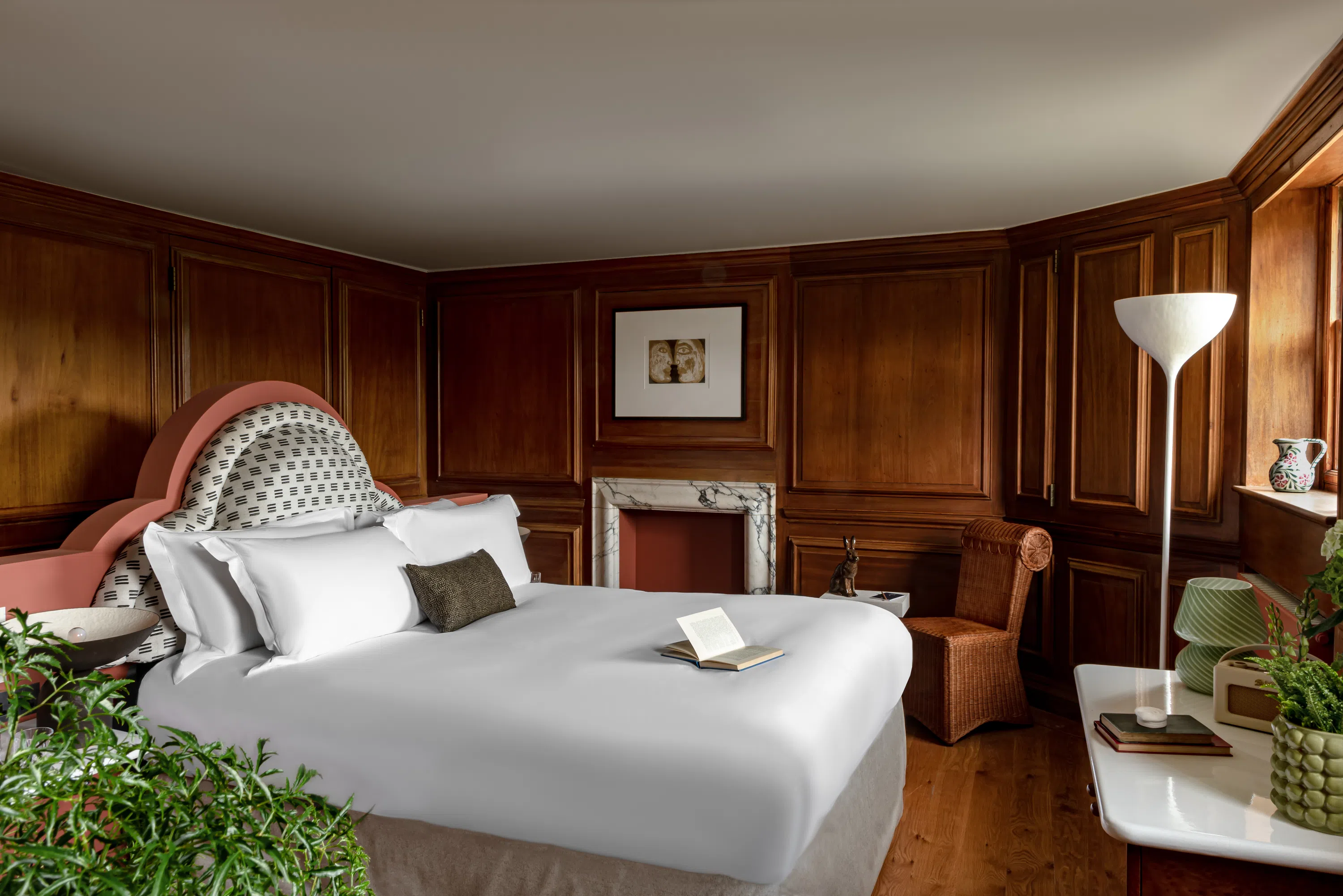 Hotel room with a large bed, wooden panel walls, wicker chair, floor lamp, and plants near a window.