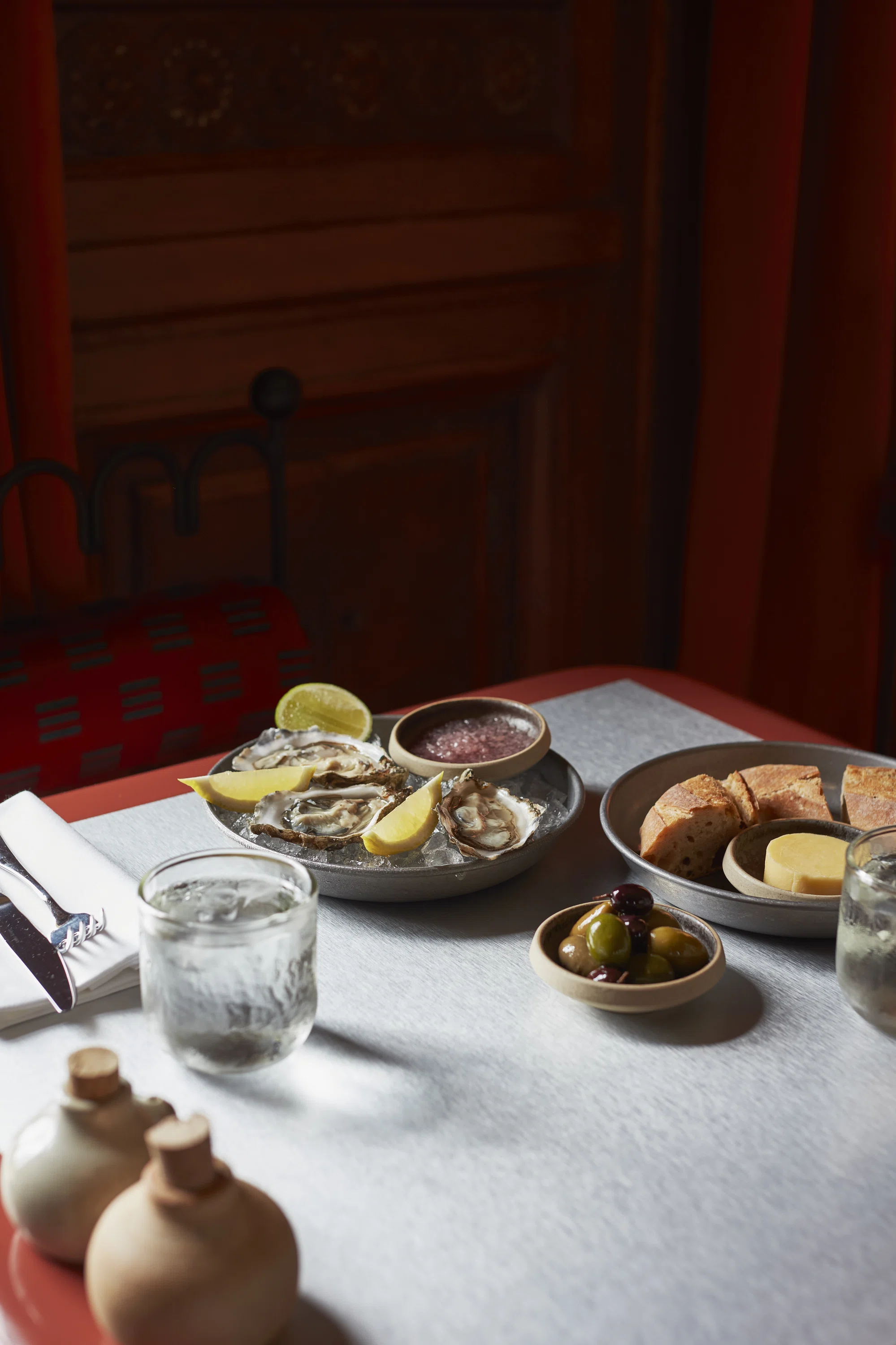 Table with oysters on ice, lemon wedges, olives, bread, butter, and glasses of water.