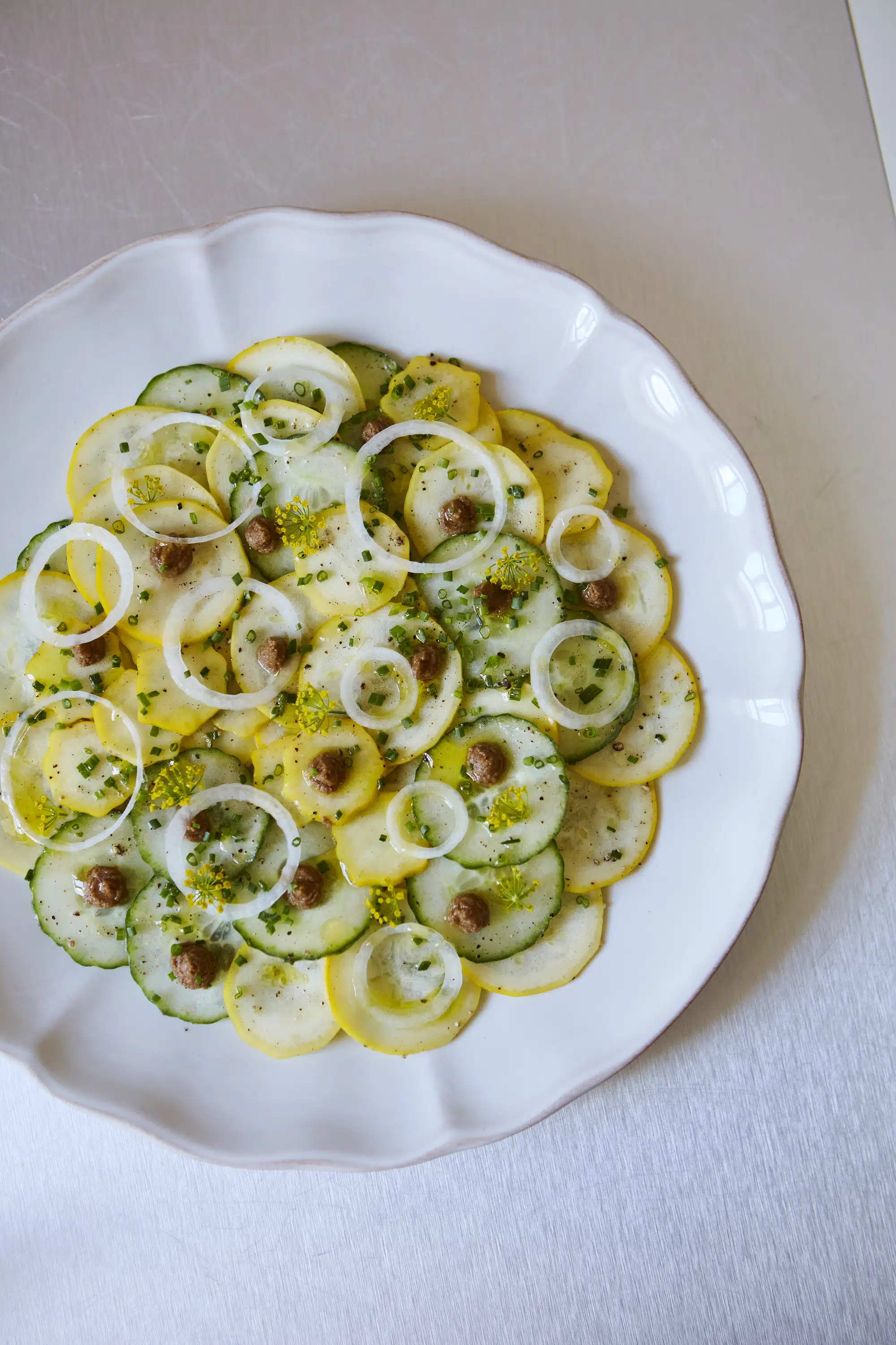 White plate with sliced cucumber, yellow squash, onion rings, chives, and small dollops of brown spread.