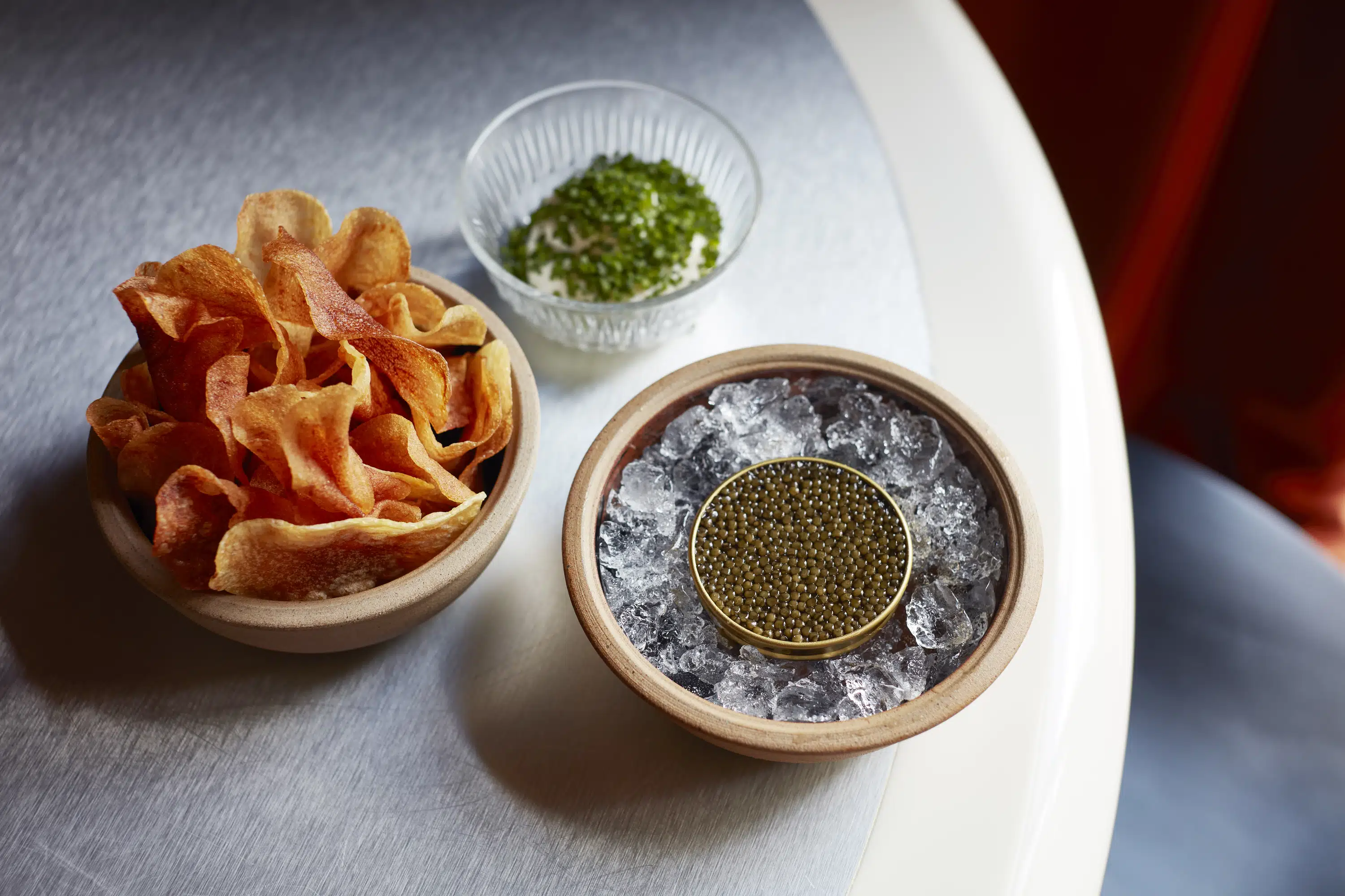 Bowl of potato crisps with a small bowl of caviar on ice and a glass bowl with cream and herbs.