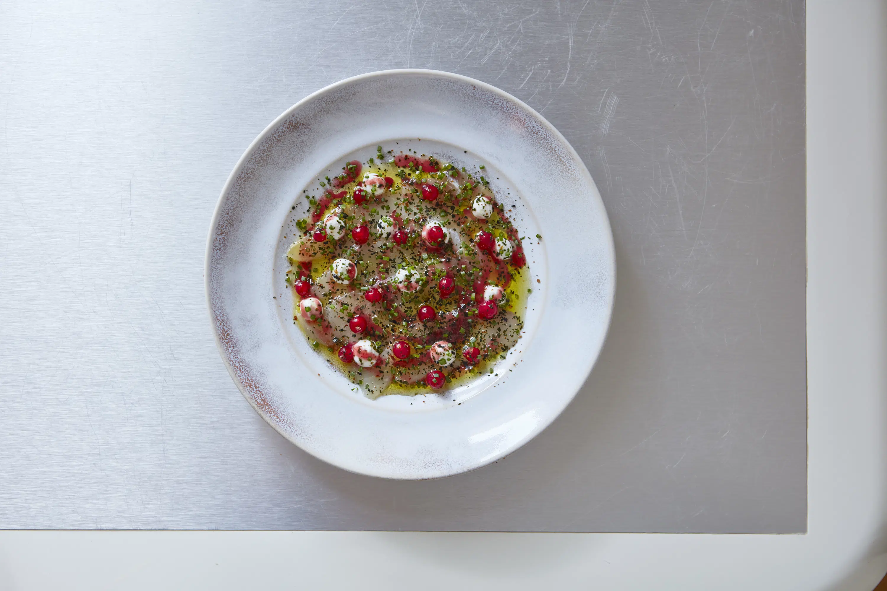 White plate with thinly sliced fish, red berries, creamy dots, herbs, and oil on a metal surface.