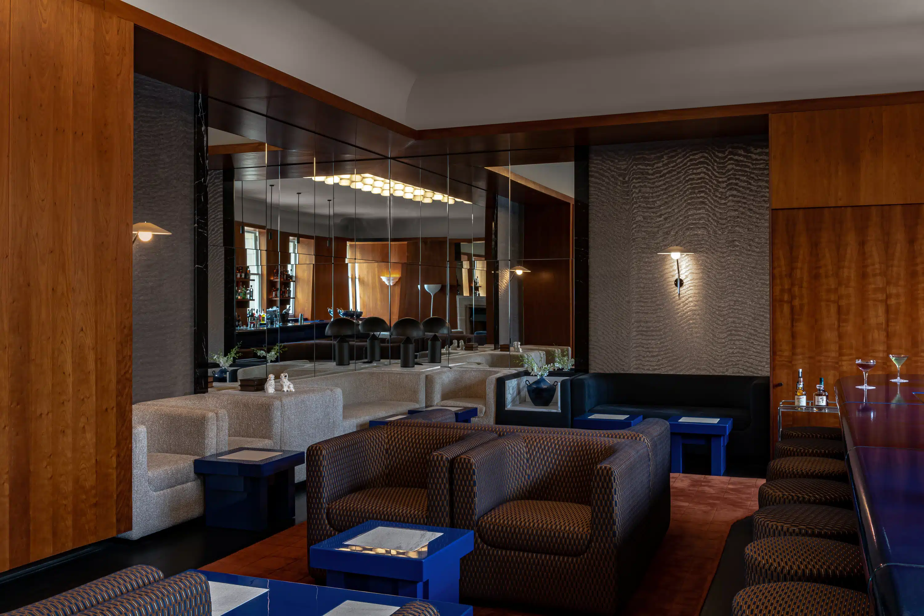 Modern hotel bar with patterned armchairs, blue tables, mirrored wall, and a row of bar stools with drinks.