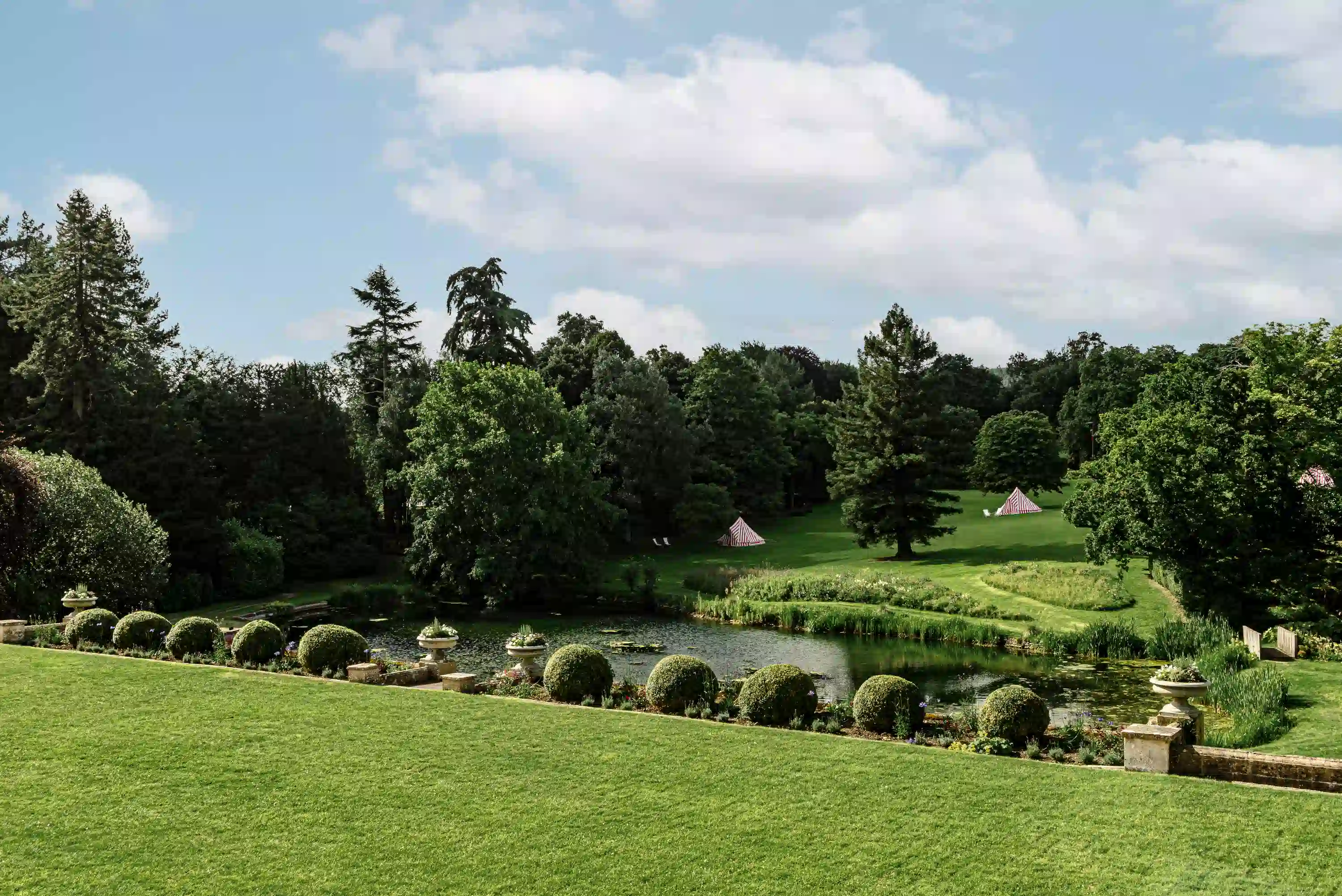 Manicured garden with trimmed bushes, a pond, tall trees, and striped tents on a sunny day.