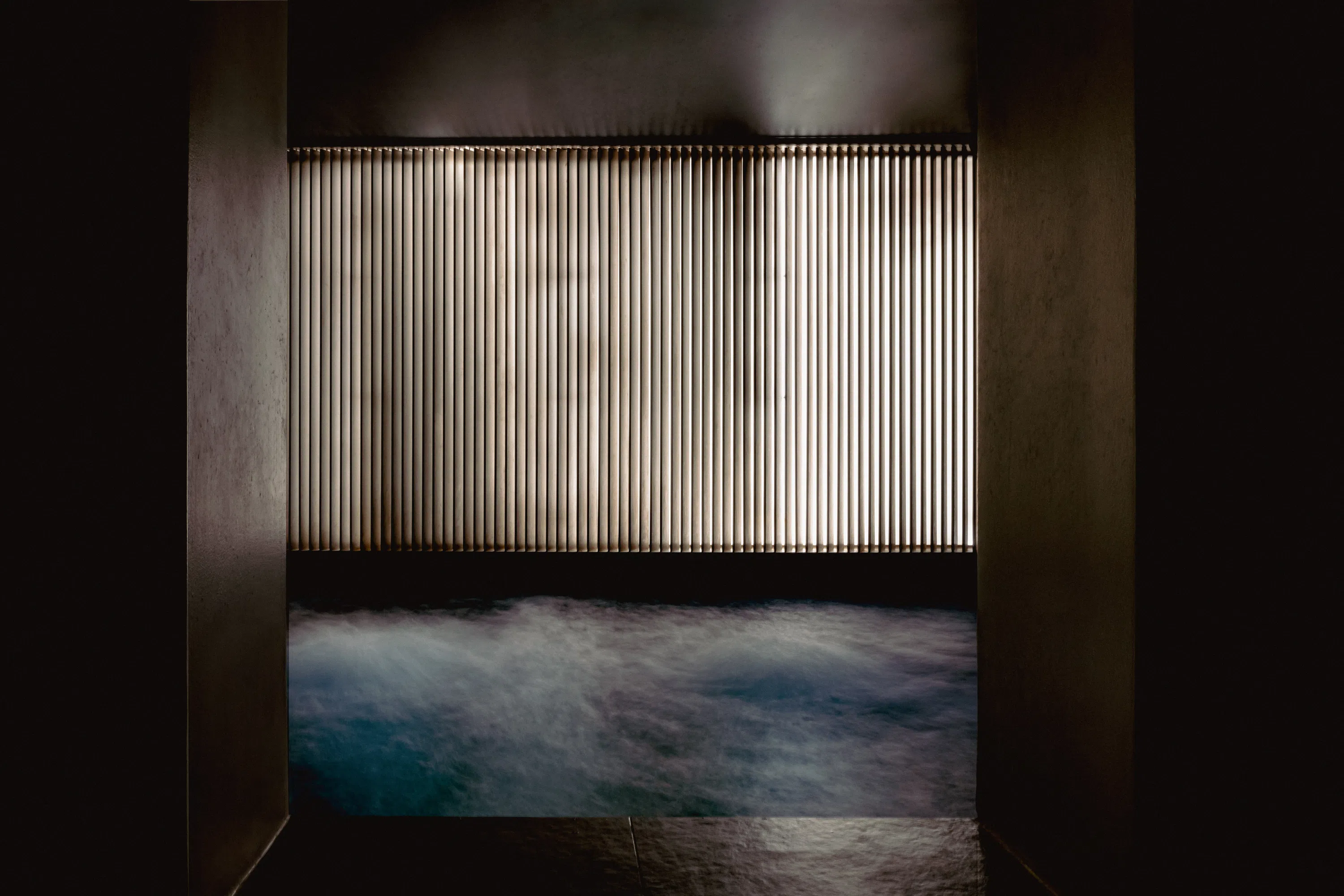 Dimly lit indoor pool or spa with bubbling water and vertical slatted window in background.