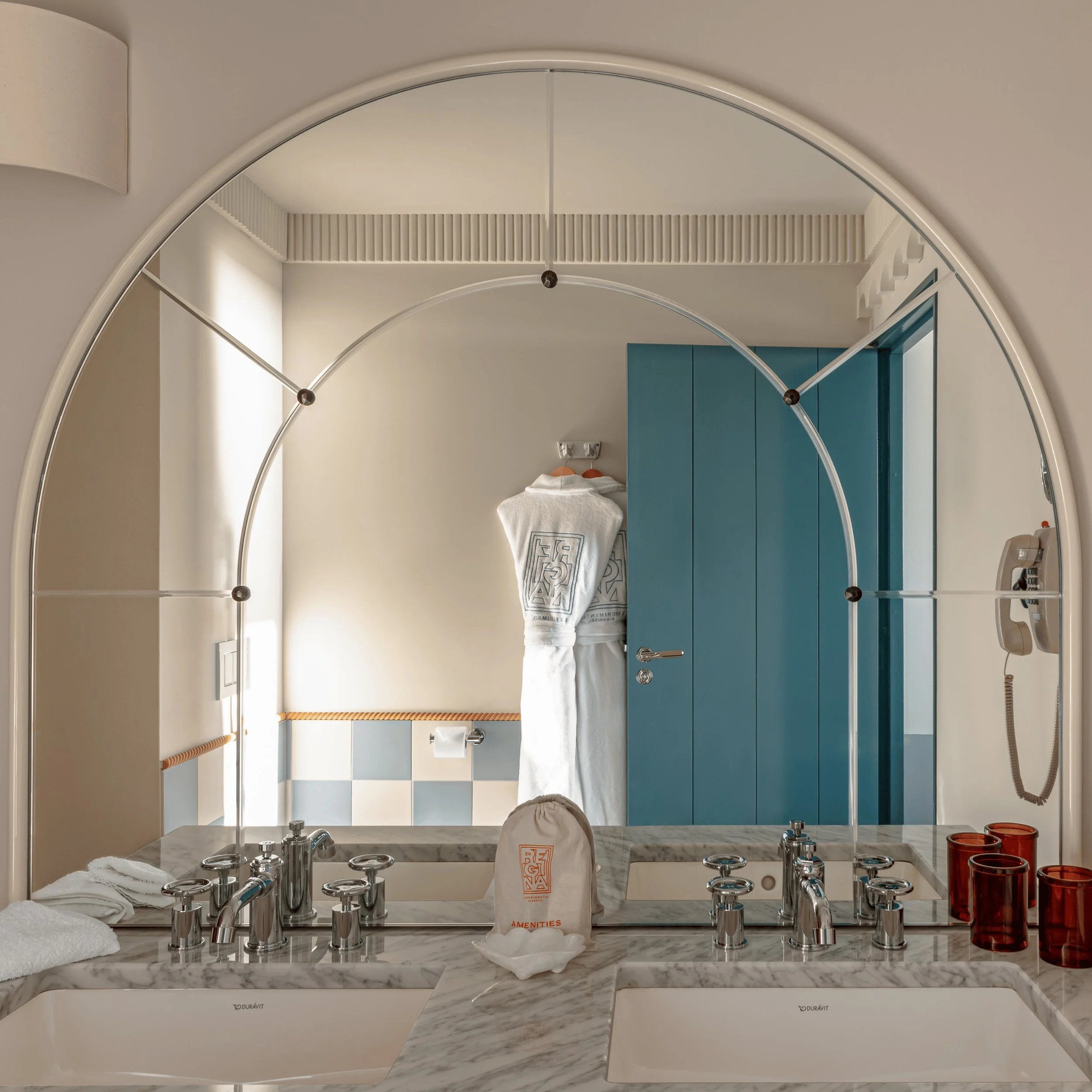 Modern bathroom with double marble sink, blue door, white bathrobe, and amber glass cups.