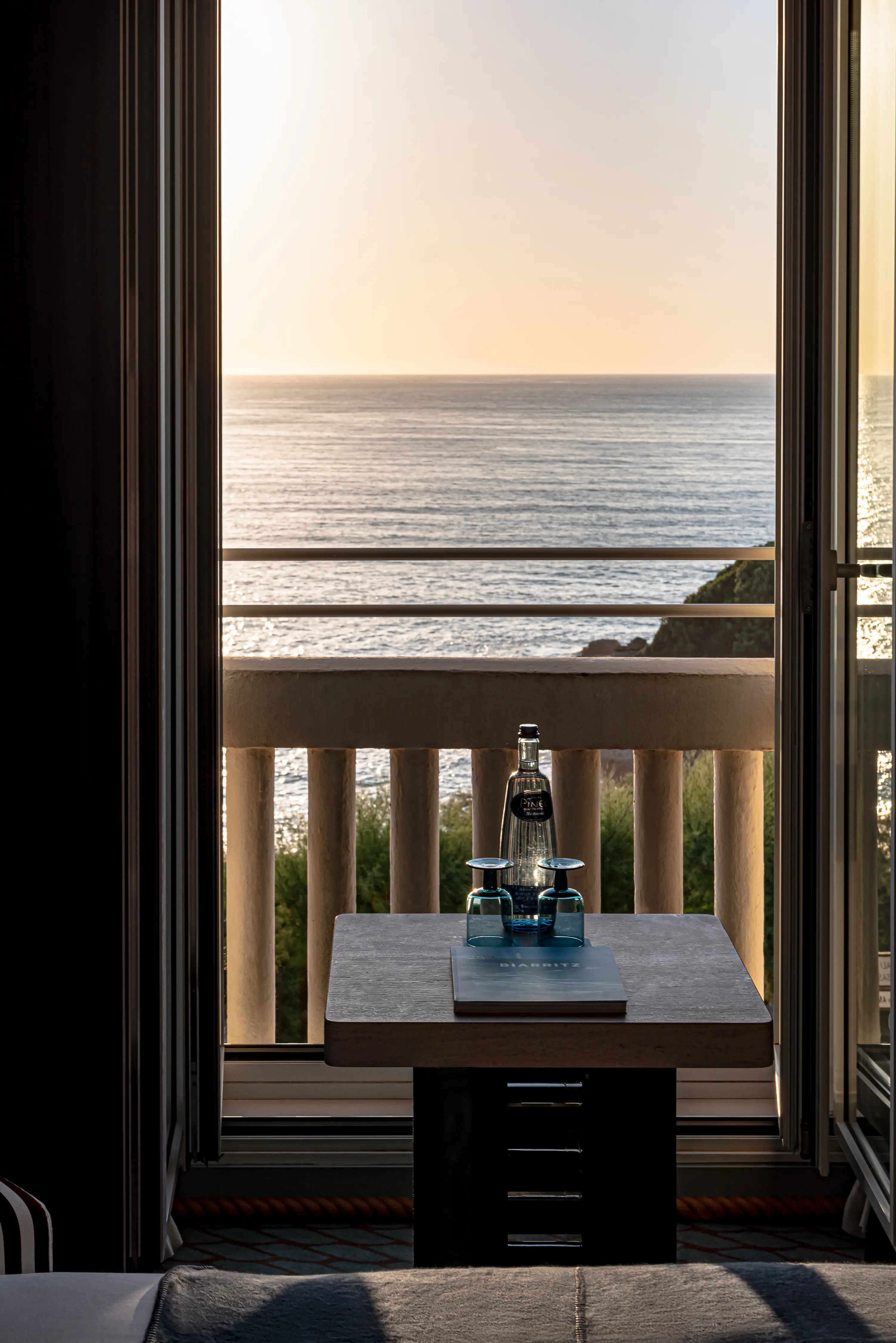 Table with two blue glasses and a bottle by an open balcony overlooking the sea at sunset.