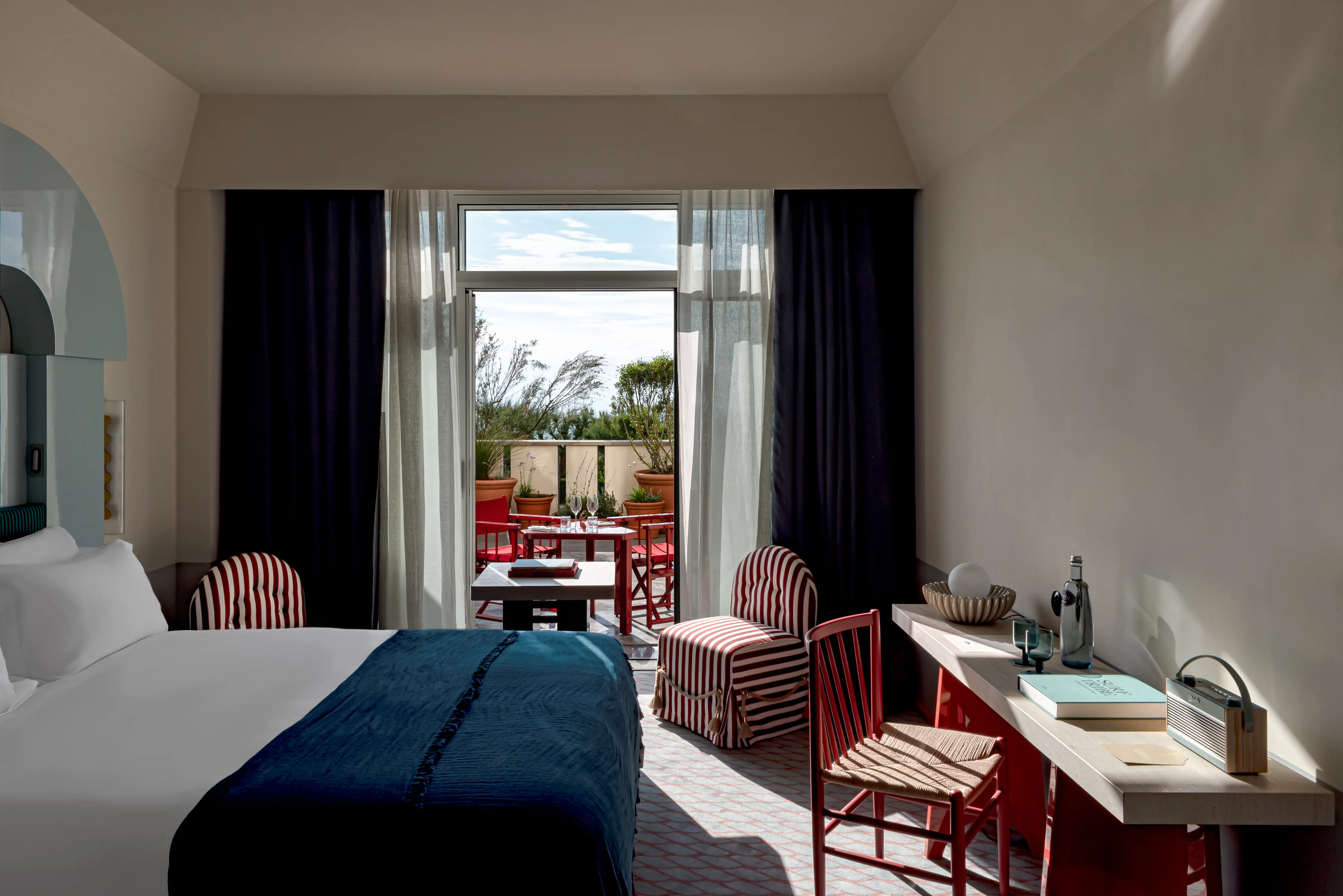 Hotel room with a bed, striped chairs, a desk, and a balcony with red furniture and plants outside.