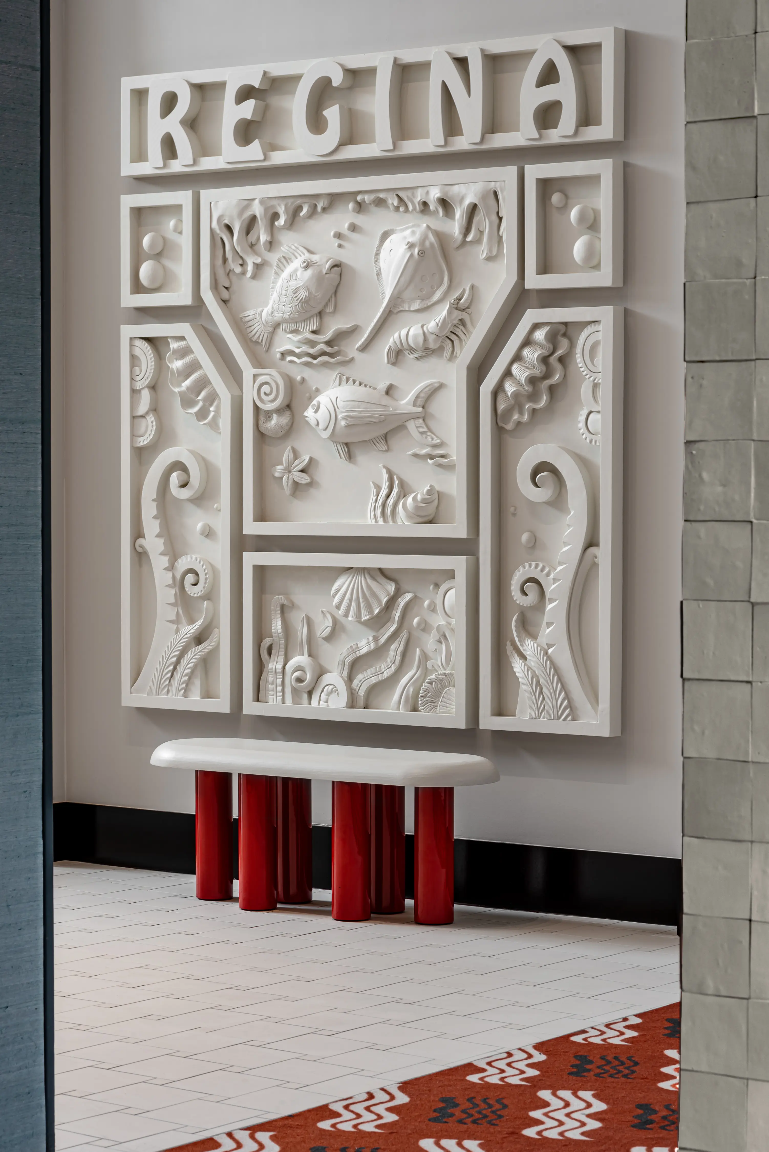 White wall art with ocean-themed reliefs and the word REGINA above a white bench with red legs.