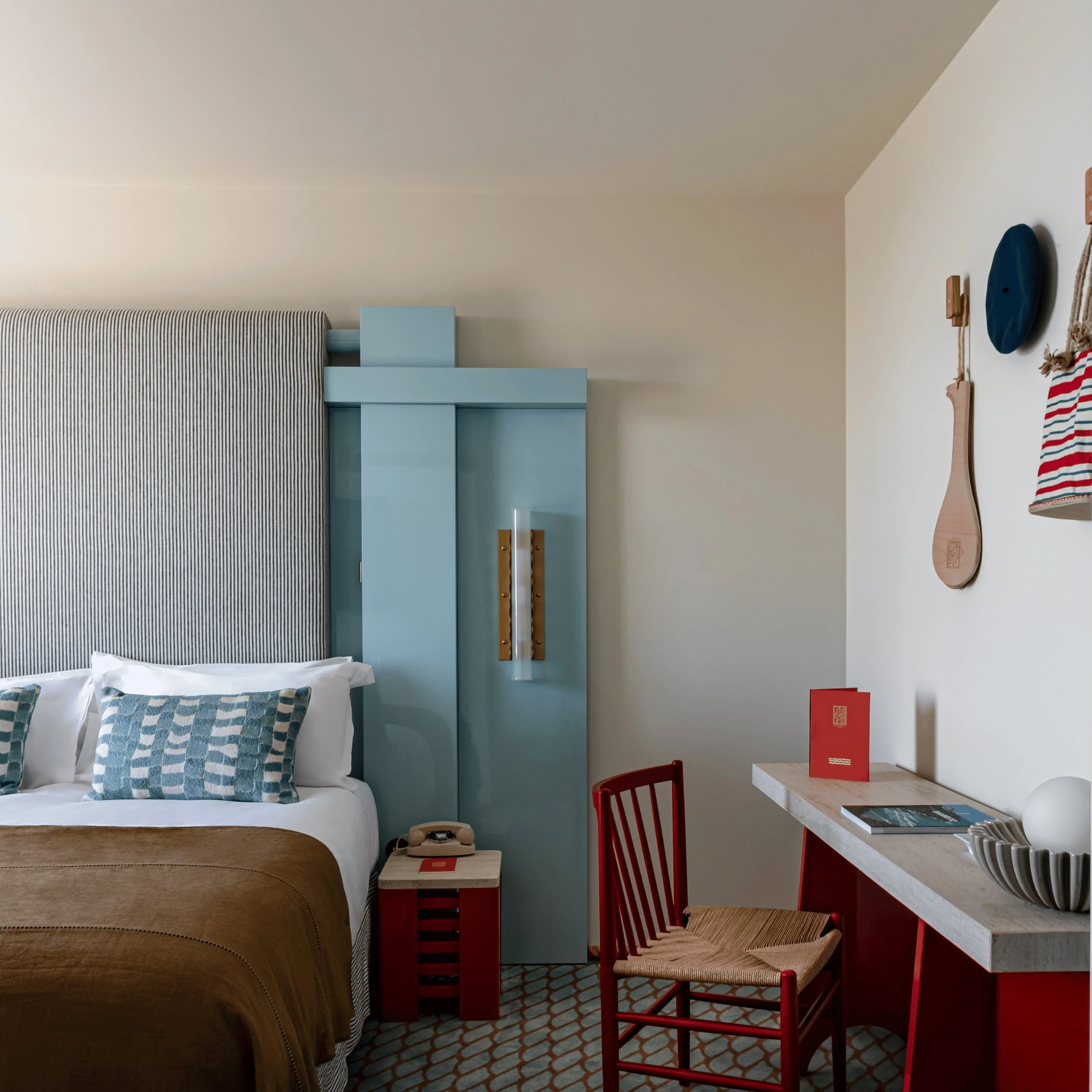 Hotel bedroom with bed, blue headboard, brown blanket, red chair, and desk with decor and books.