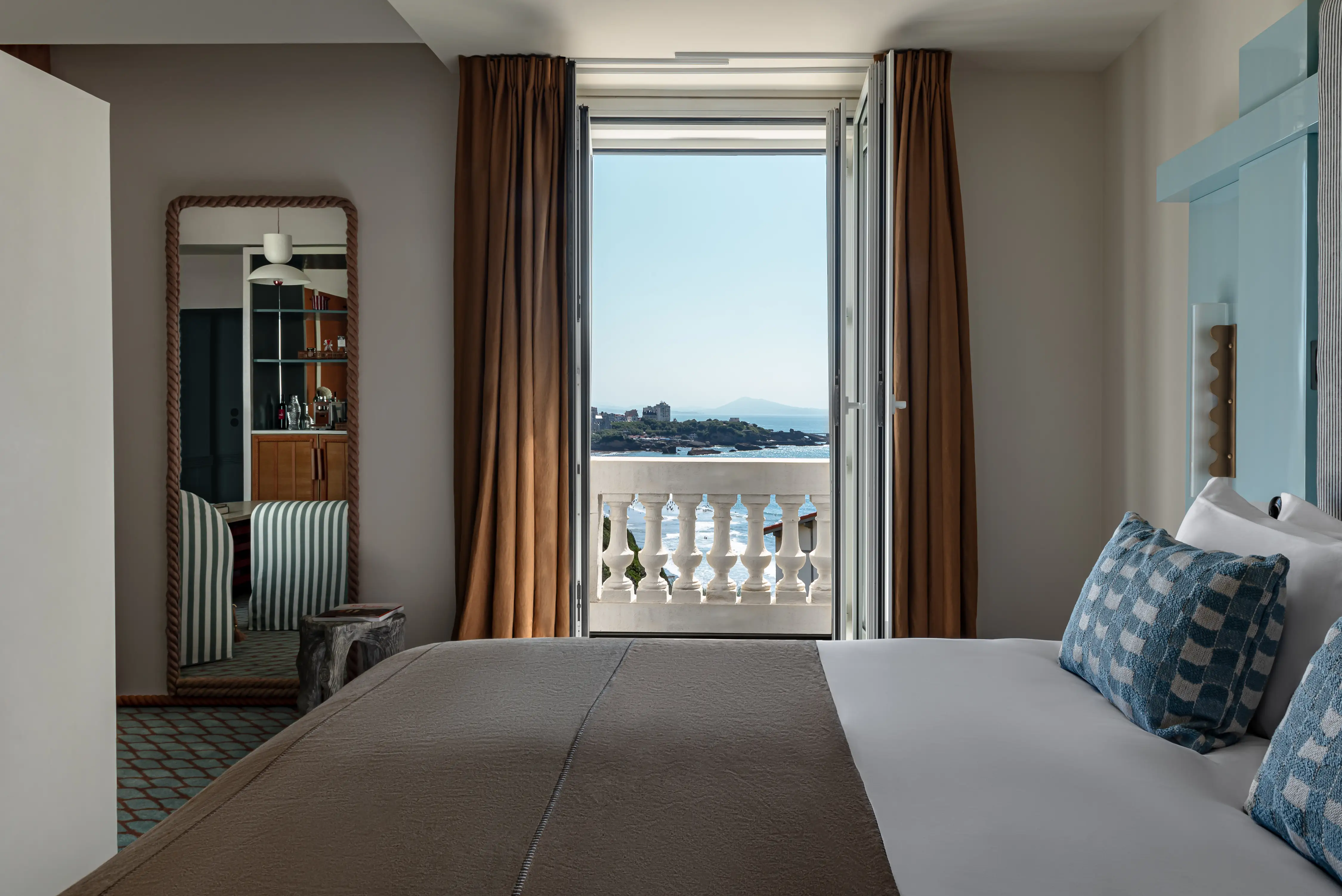 Hotel room with bed, striped pillows, large mirror, and open door showing sea view and balcony.