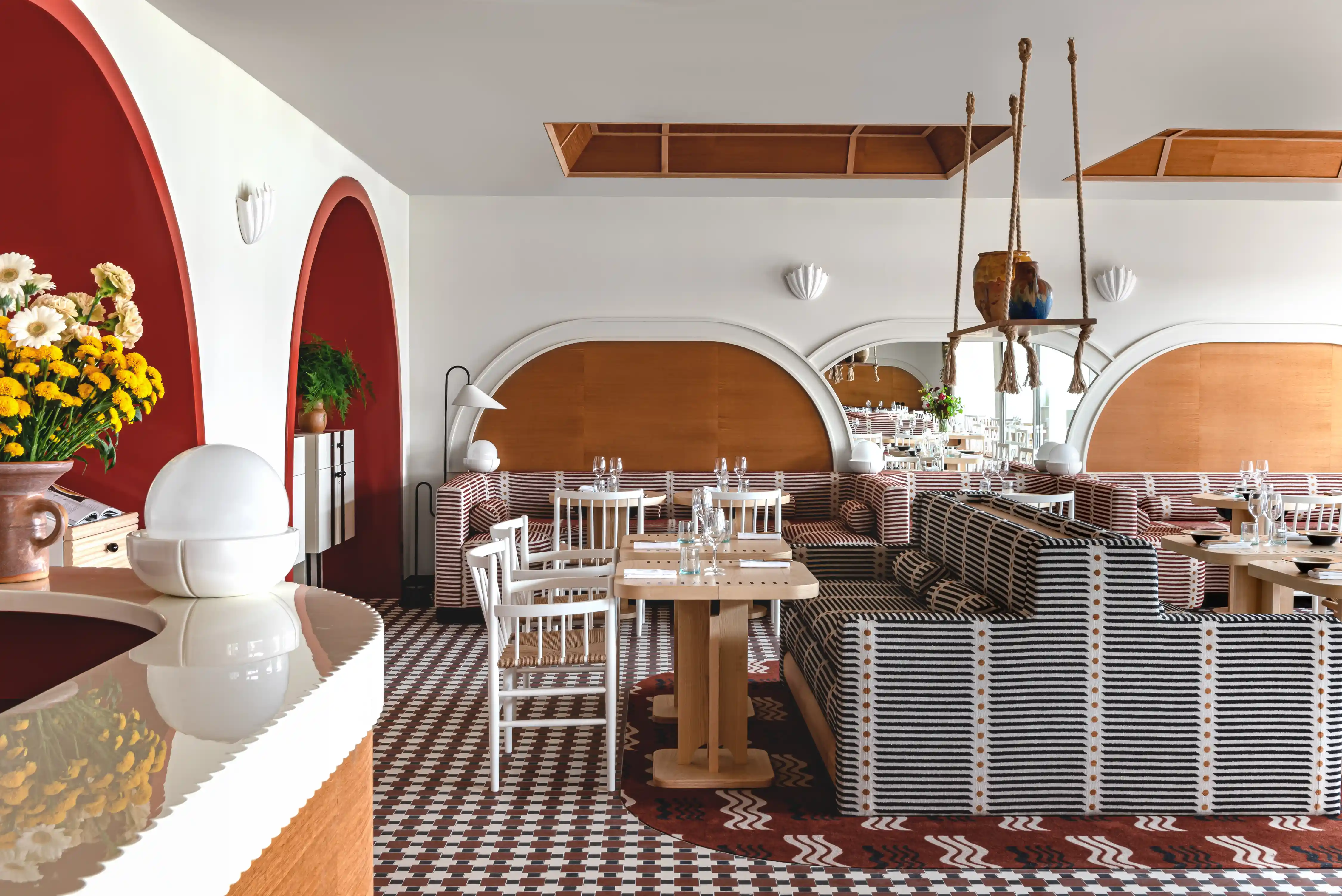Modern restaurant interior with striped sofas, wooden tables, white chairs, and red arched wall accents.