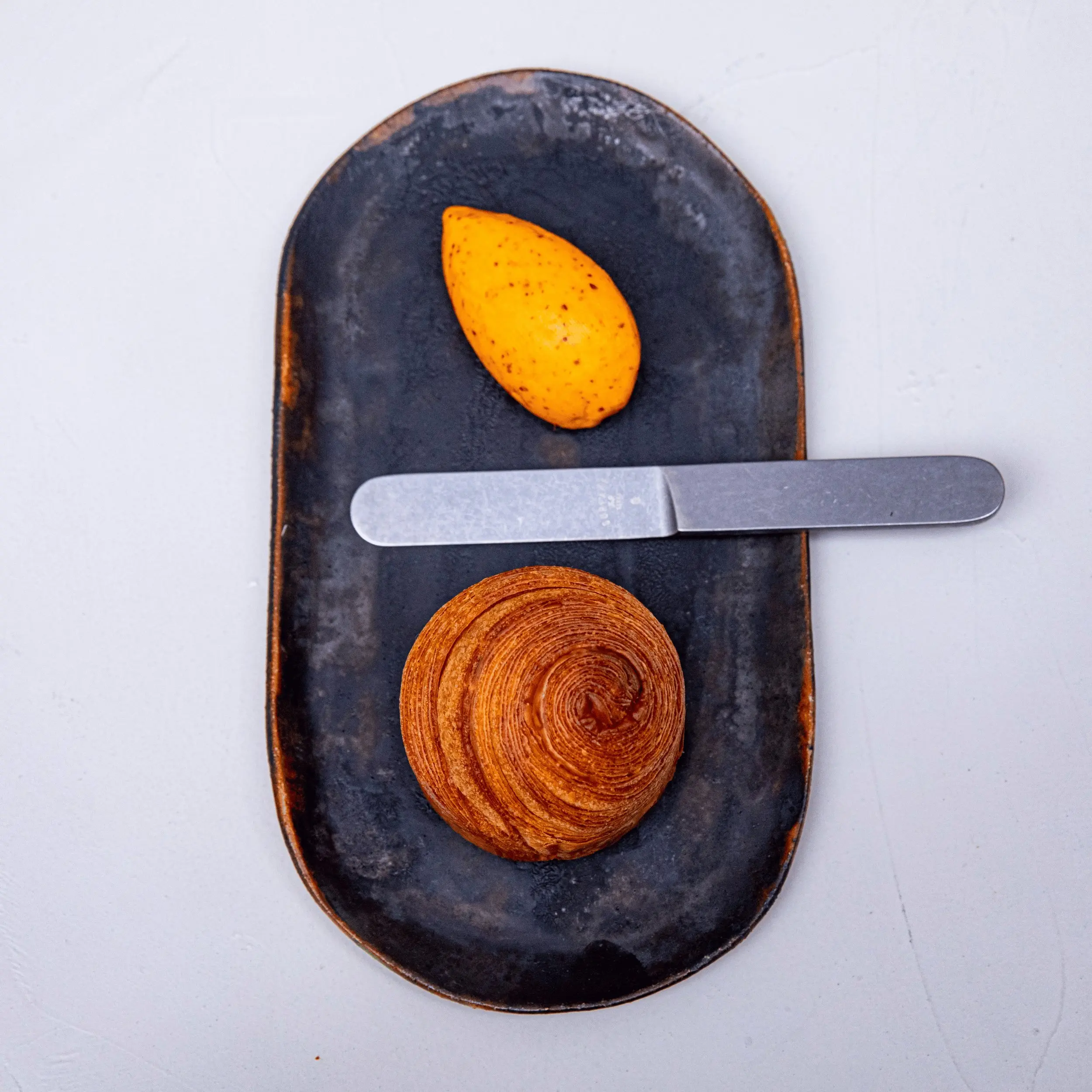 Butter pat, metal butter knife, and round croissant on black oval plate against white background.