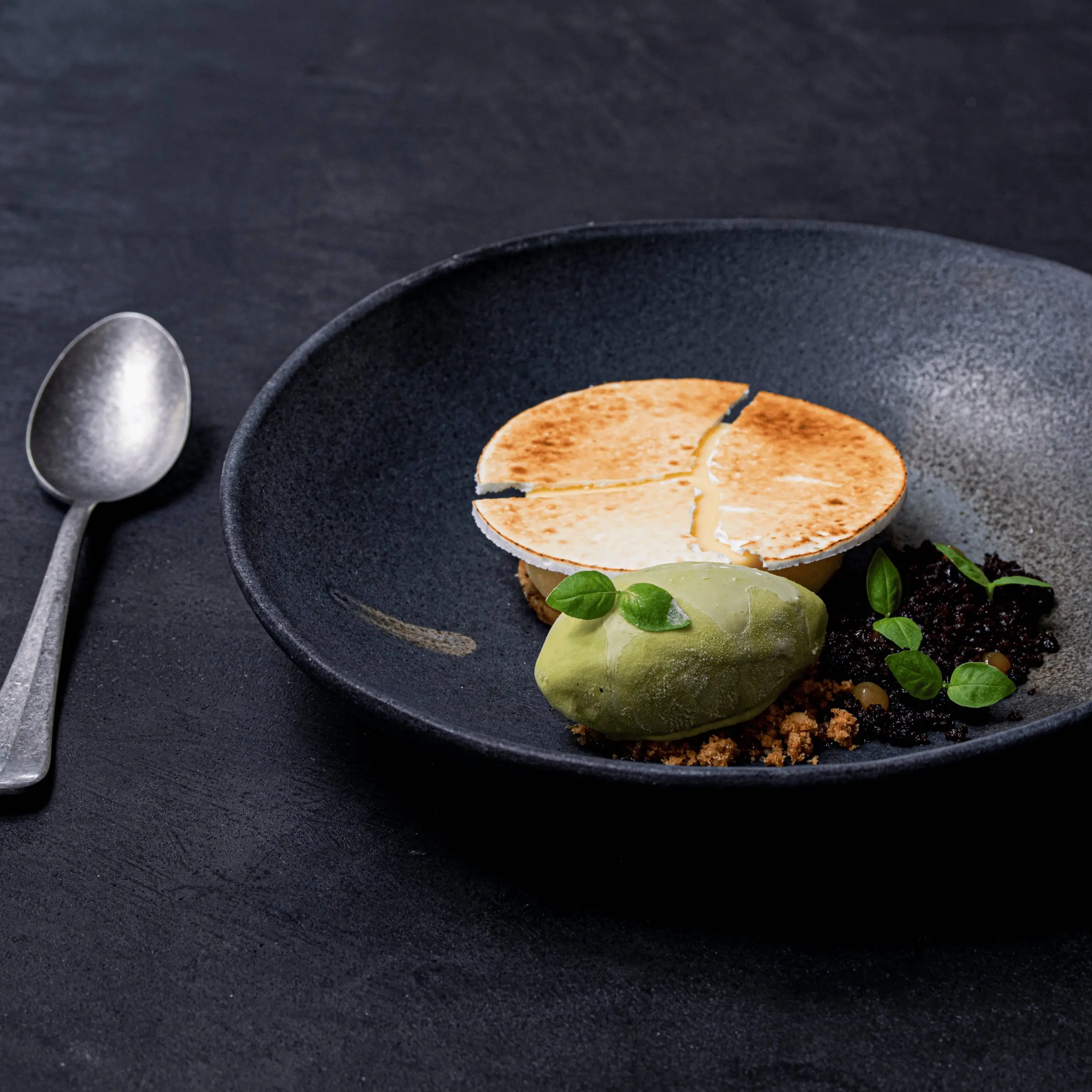 Gourmet dessert with a toasted round top, green sorbet, crumbles, and herbs on a black plate.