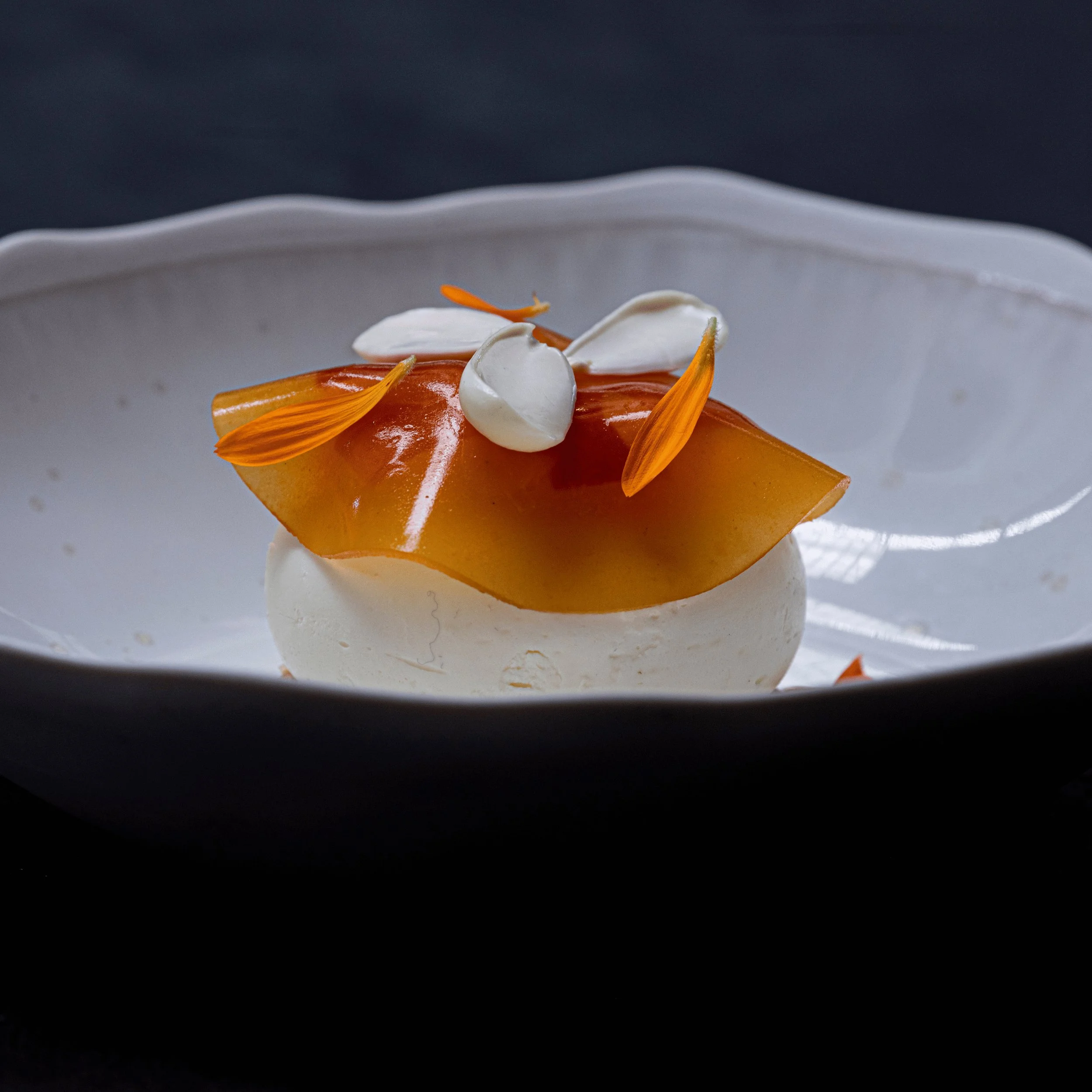 Elegant dessert with translucent amber sheet, white cream, and orange flower petals on white plate.