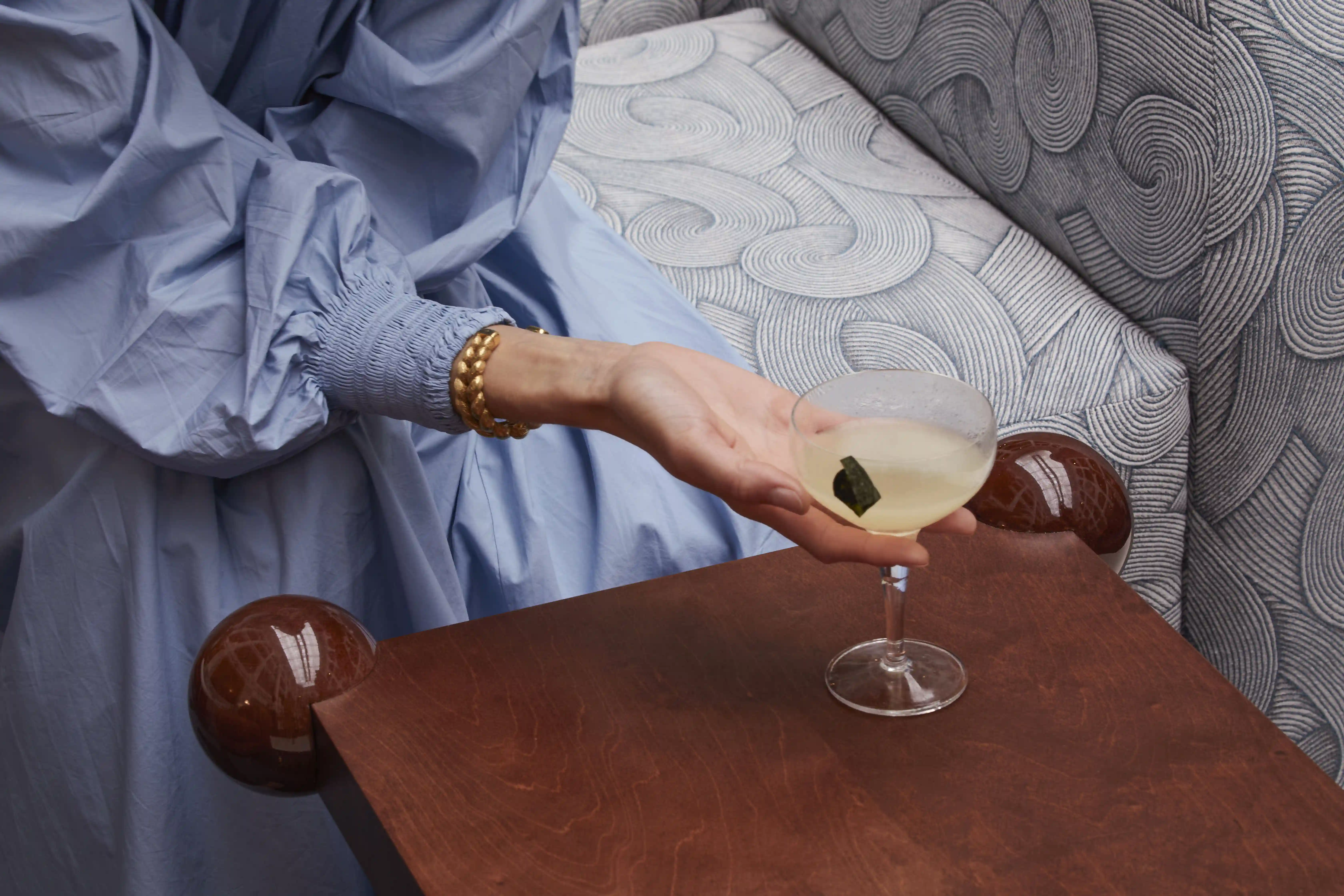 Hand with chunky gold bracelet holding a coupe glass with a light cocktail and garnish.