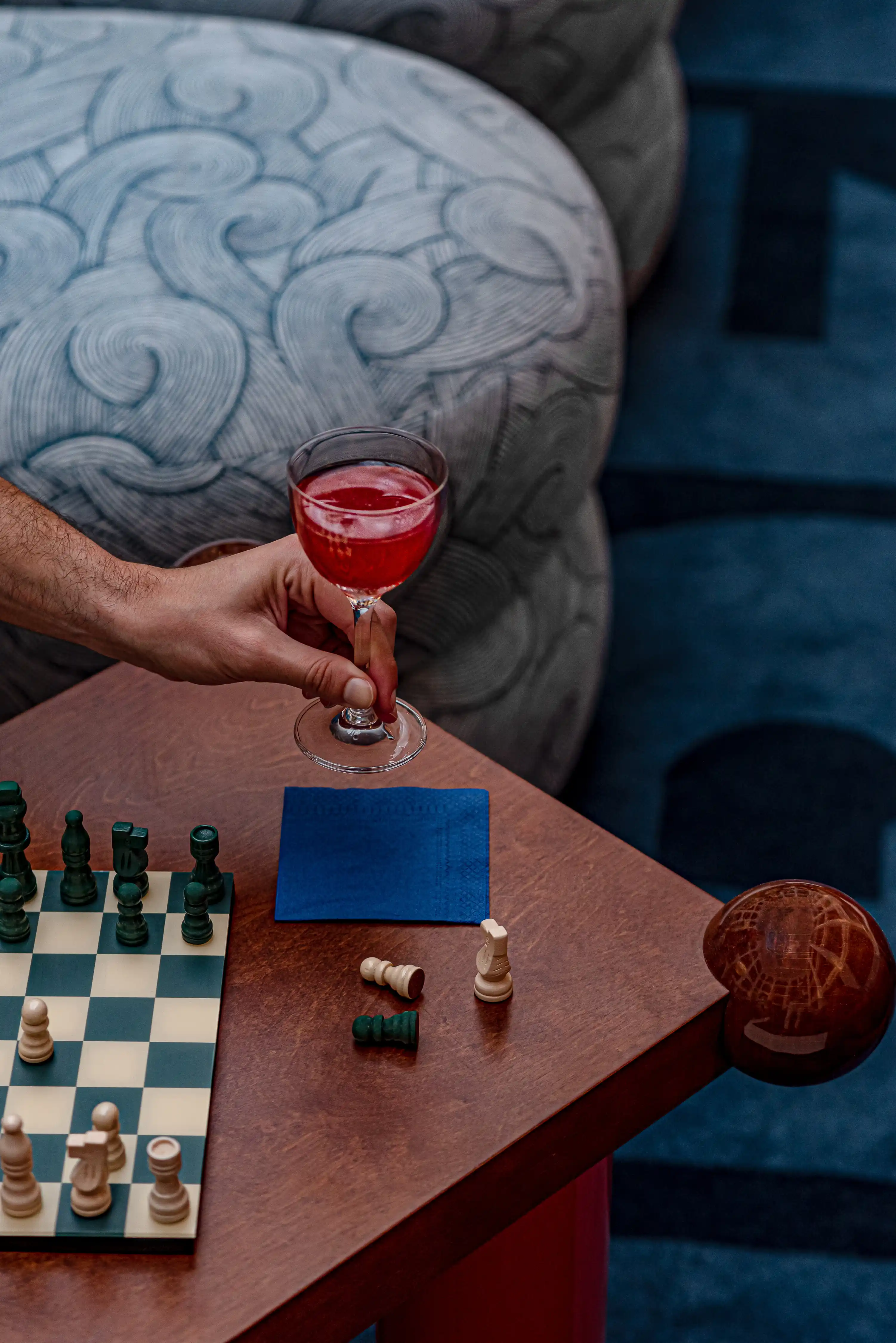 Hand holding a red cocktail glass above a table with a chessboard and fallen chess pieces.