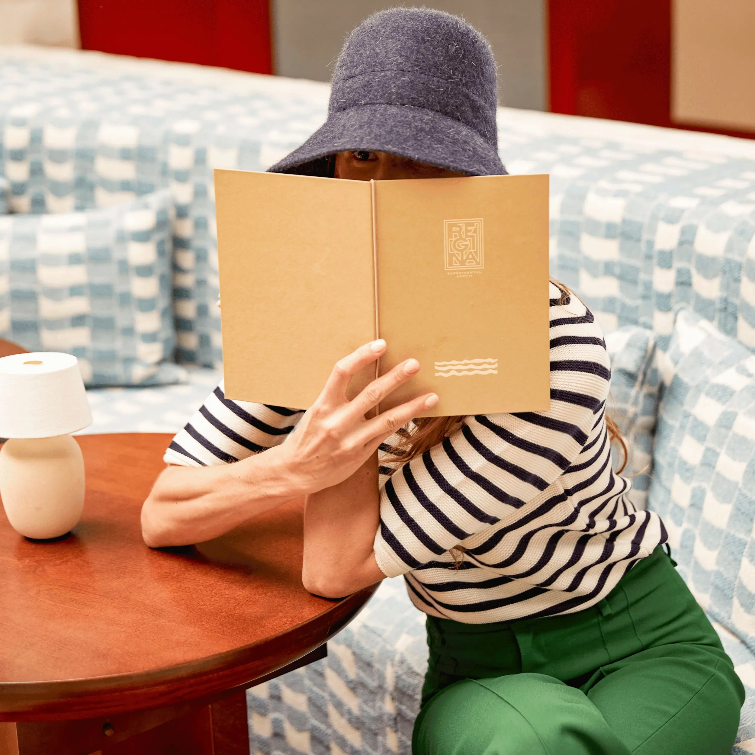 Person in striped shirt and blue hat sitting on patterned couch, holding a brown menu.