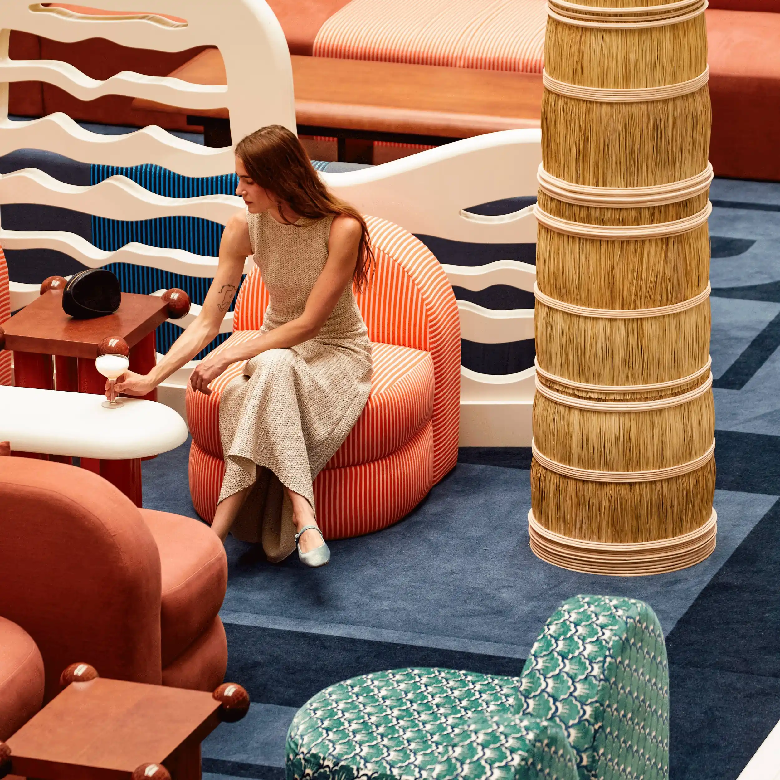 Woman in beige dress sitting on striped orange chair holding a cocktail glass in a colorful lounge area.