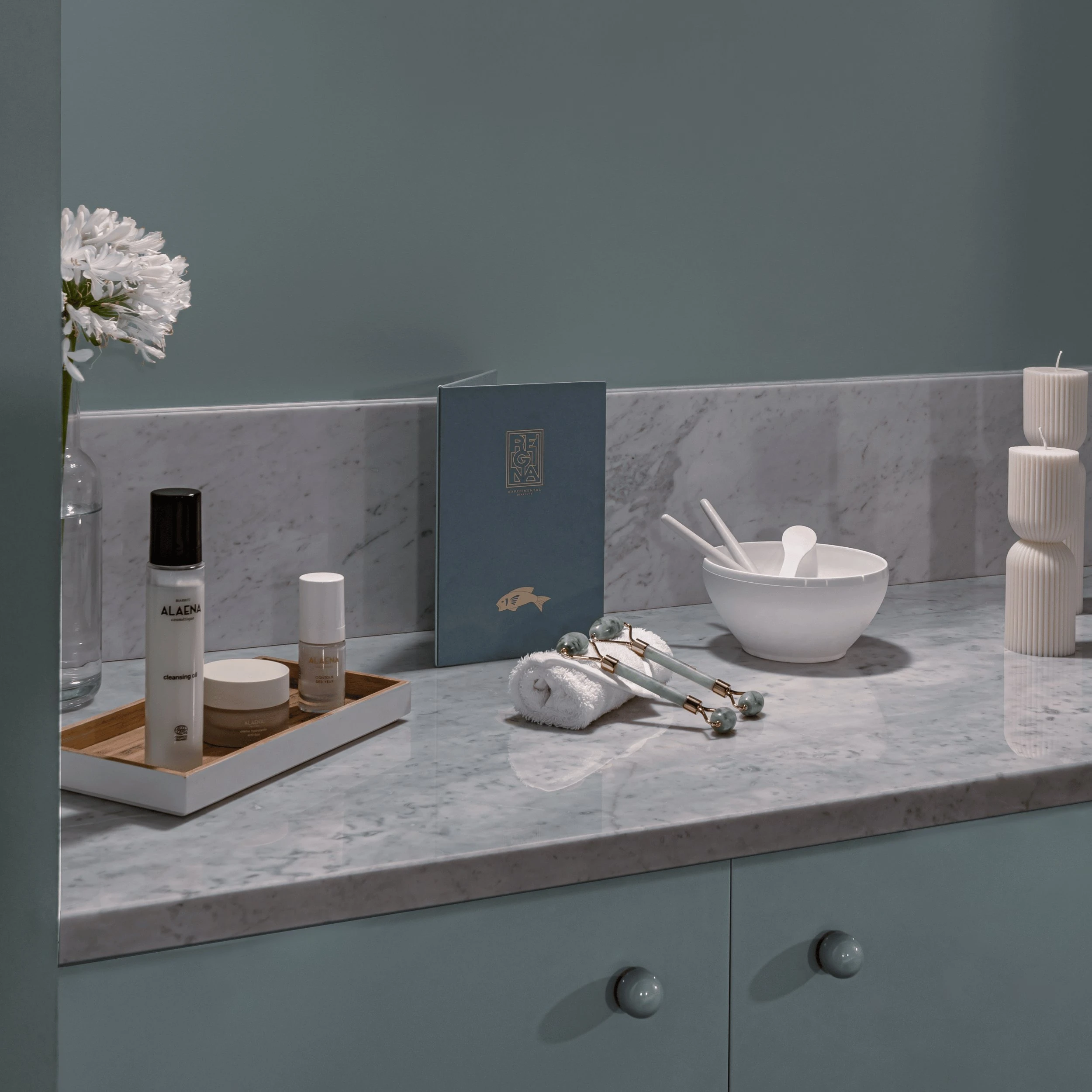 Marble countertop with skincare products, facial rollers, a bowl with spoons, candles, and a flower in a vase.