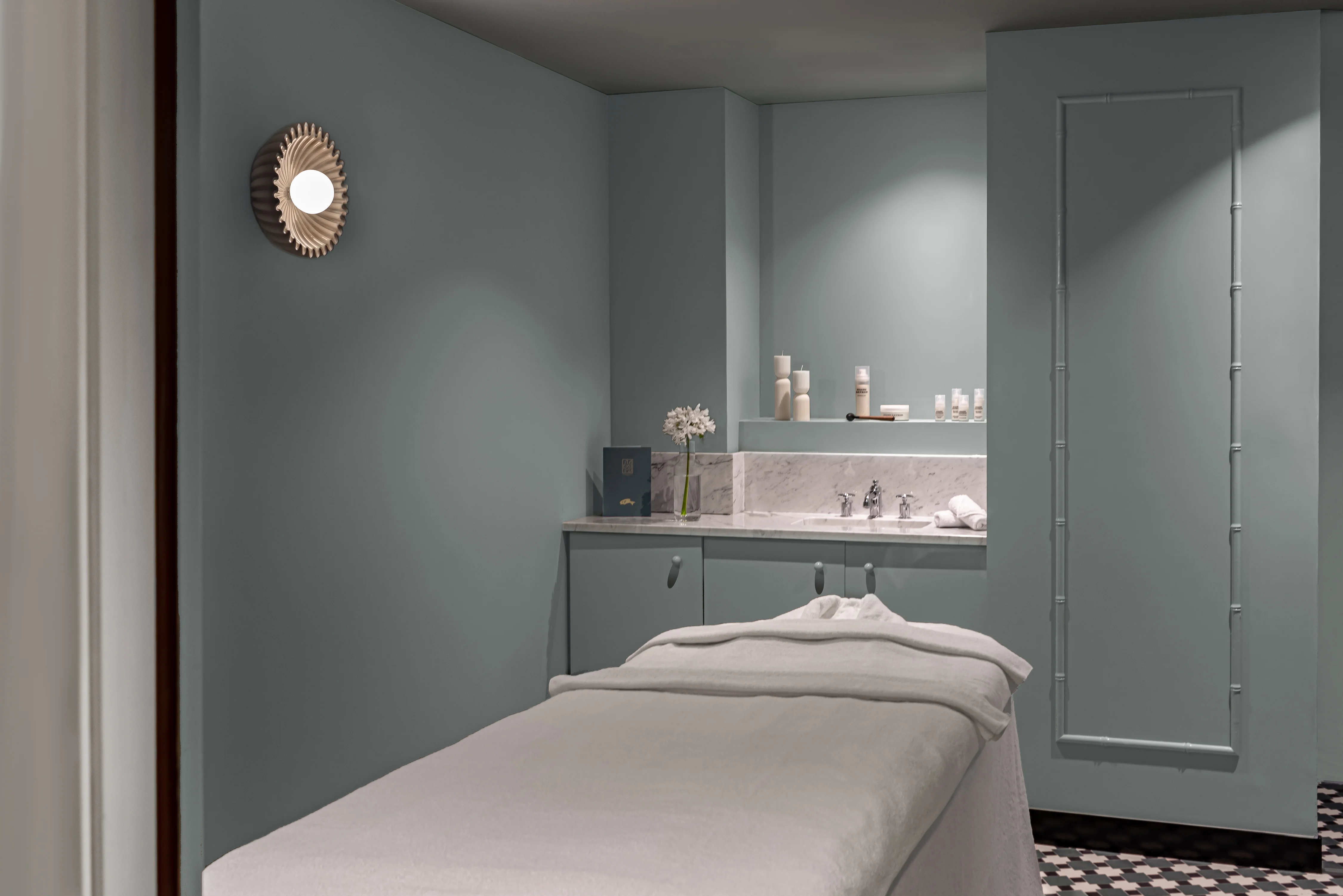 Spa treatment room with massage table, soft gray walls, wall light, and skincare products on marble counter.