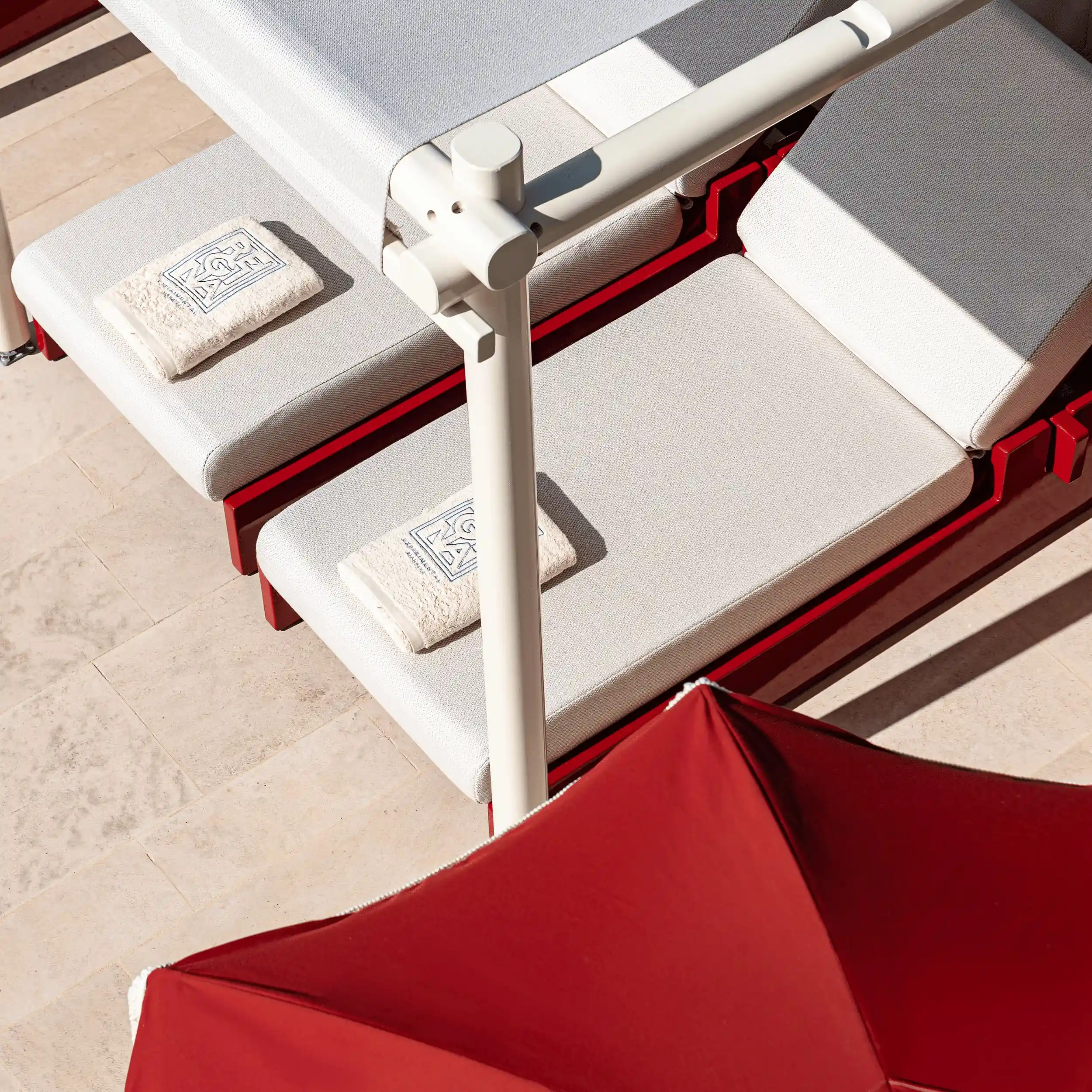 Two white cushioned lounge chairs with folded towels, beside a red patio umbrella on a tiled floor.