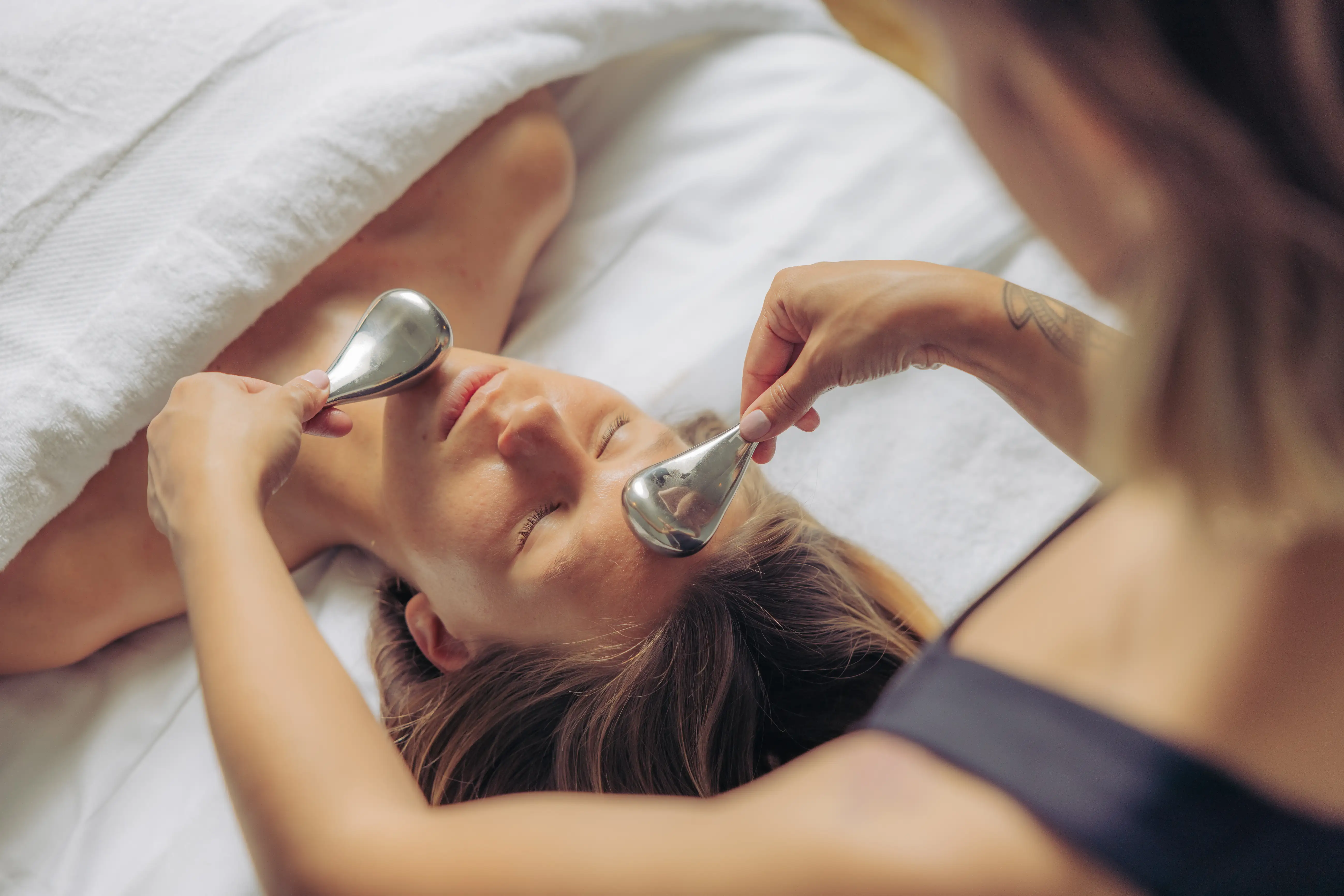 Person receiving a facial massage with metal tools while lying under a white towel.
